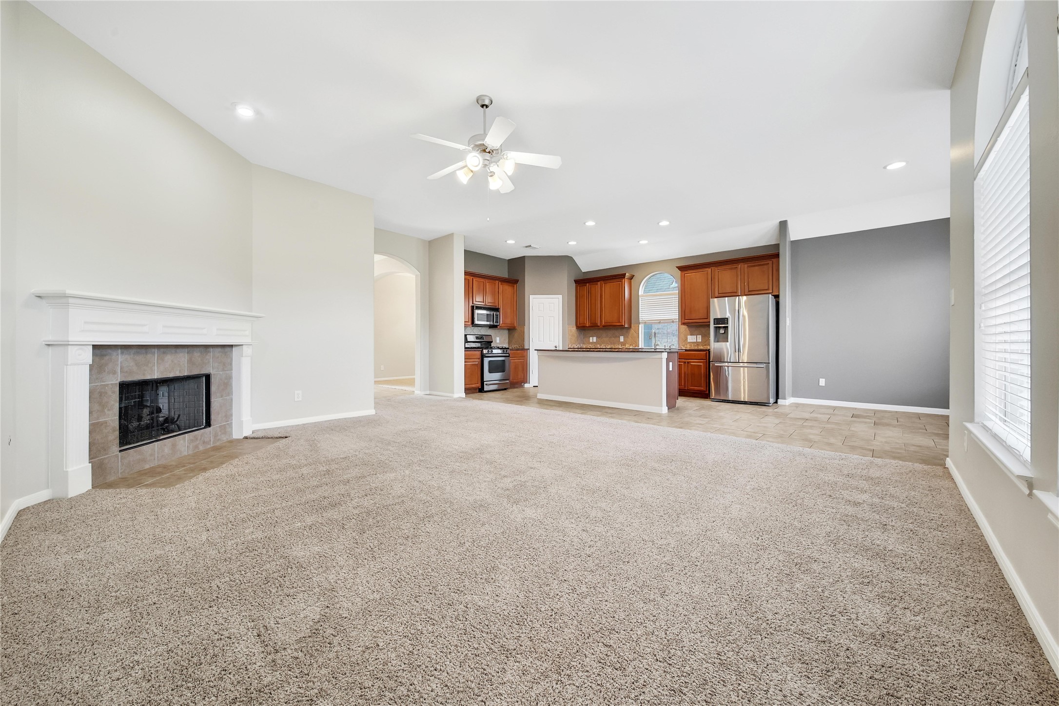 This is a spacious, open-concept living room with plush carpeting, featuring a cozy fireplace and a seamless transition to a tiled kitchen area with wood cabinets and stainless steel appliances. The room is well-lit with natural light from large windows.