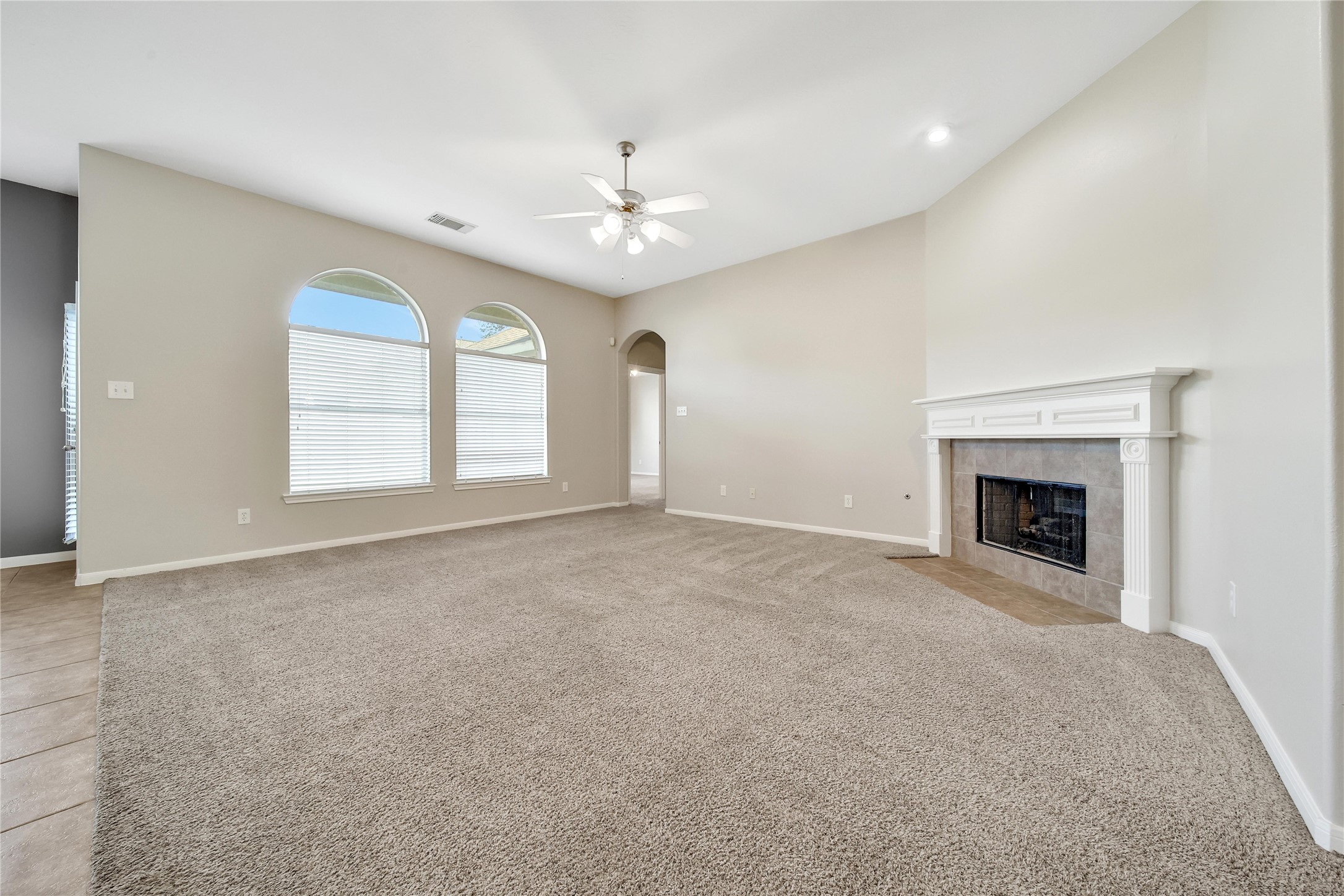 This spacious living room features plush carpeting, large arched windows for ample natural light, a ceiling fan, and an elegant fireplace as the focal point. Alcove of living room leads to primary bedroom.