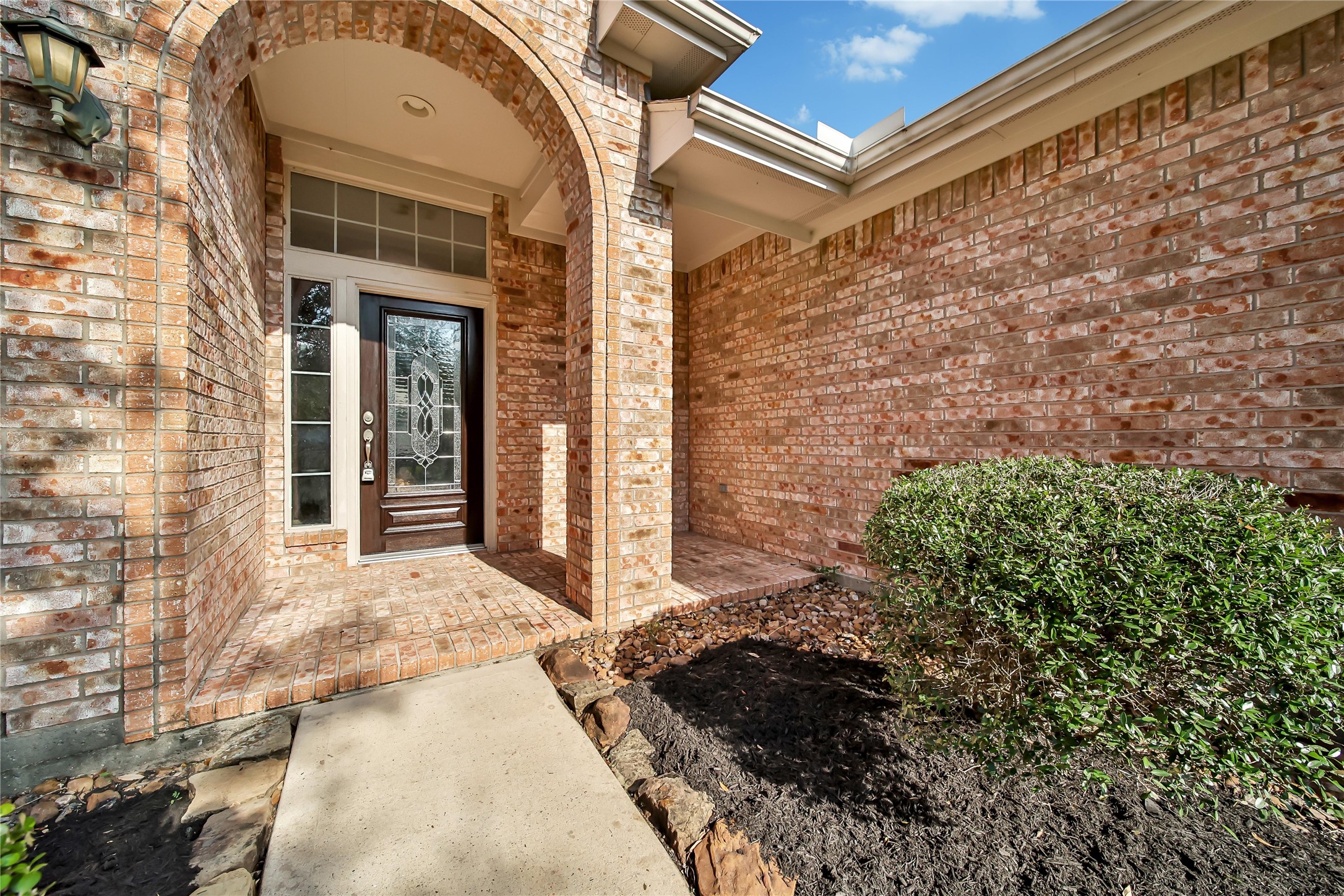This is an image of a home's welcoming entrance, featuring a brick archway leading to a wood front door with decorative glass and a sidelight. The home has a clean, well-maintained appearance with a neat landscaping touch on the side.