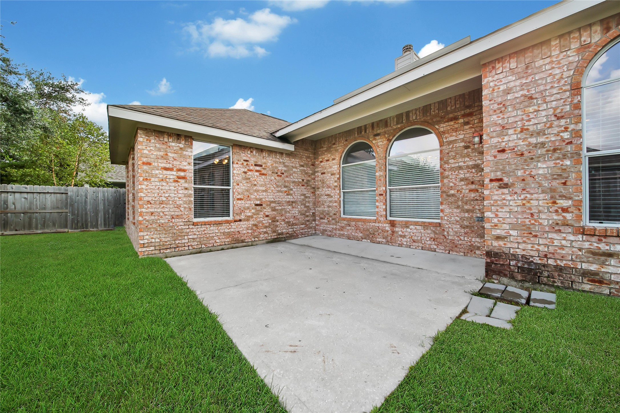 This is a well-maintained backyard with a spacious concrete patio and lawn, edged by a tall privacy fence. The exterior features a clean, classic brick finish with several windows allowing for natural light.