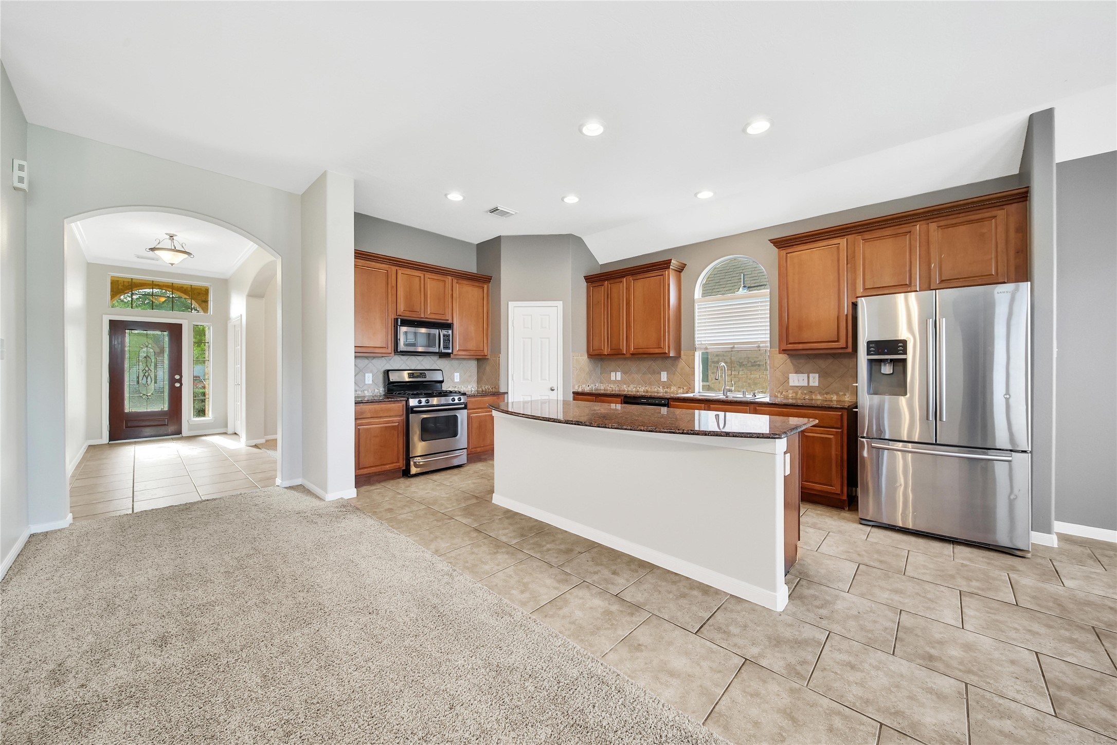 You're looking at a spacious kitchen with modern stainless steel appliances, ample wooden cabinetry, and granite countertops. The kitchen opens up to a welcoming entryway with a view of the front door through an arched passageway. The room features a blend of tile flooring in the kitchen area and plush carpeting in the adjoining space.