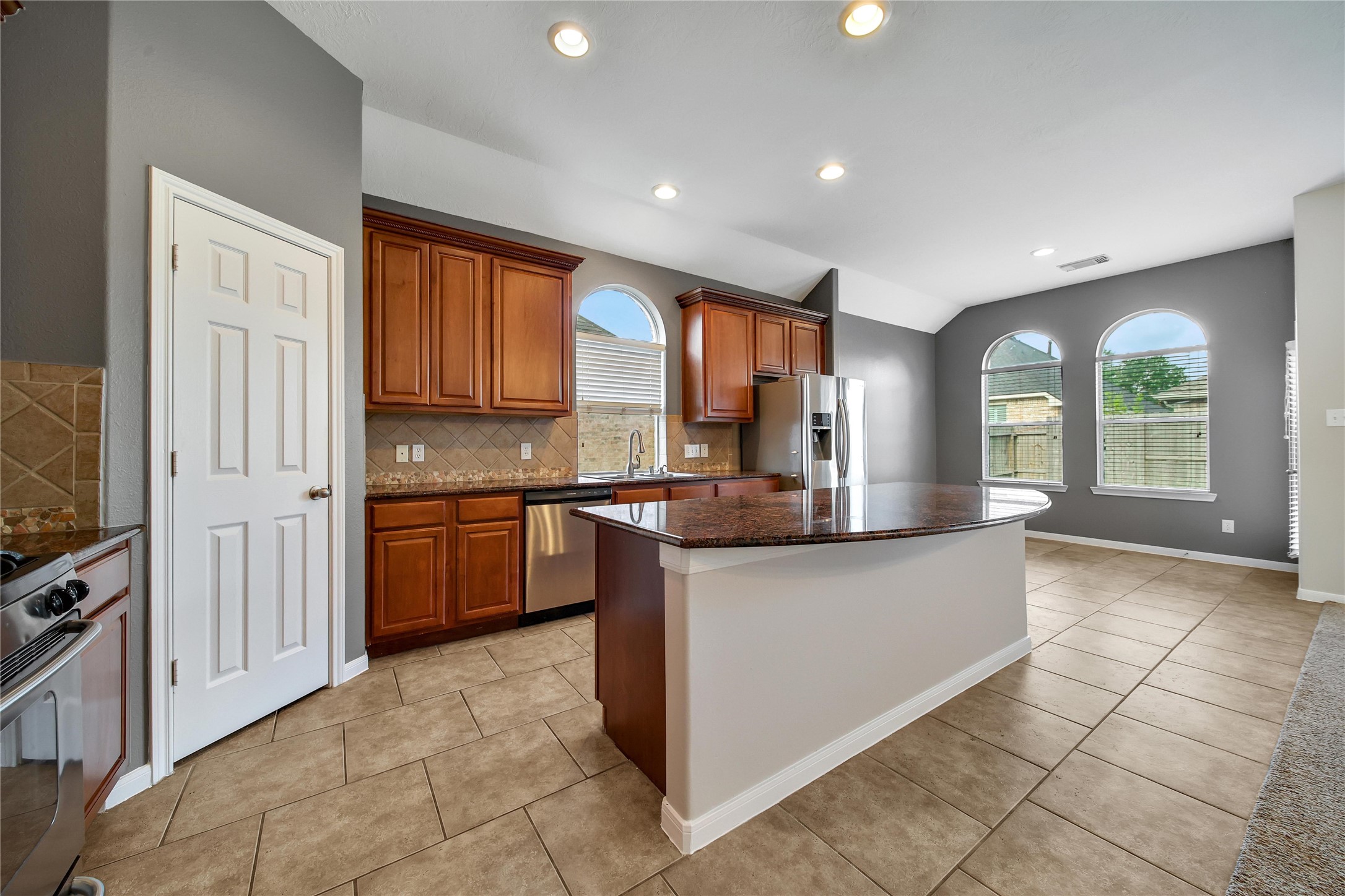 This is a spacious kitchen featuring rich wooden cabinetry, modern stainless-steel appliances, tiled flooring, and a central island with countertop space. Natural light streams in through arched windows, complementing the neutral color palette and recessed lighting.