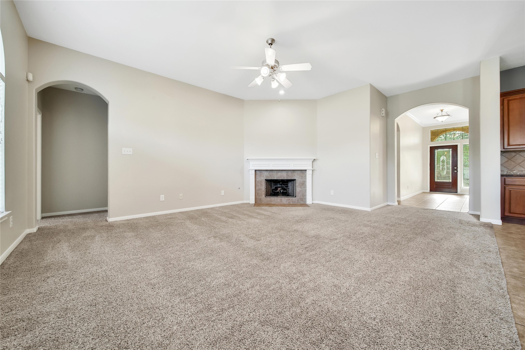 This is a spacious, carpeted living room with a fireplace and high ceilings, featuring an arched doorway leading to the entry hall and another to the adjacent room, offering a bright and airy feel.