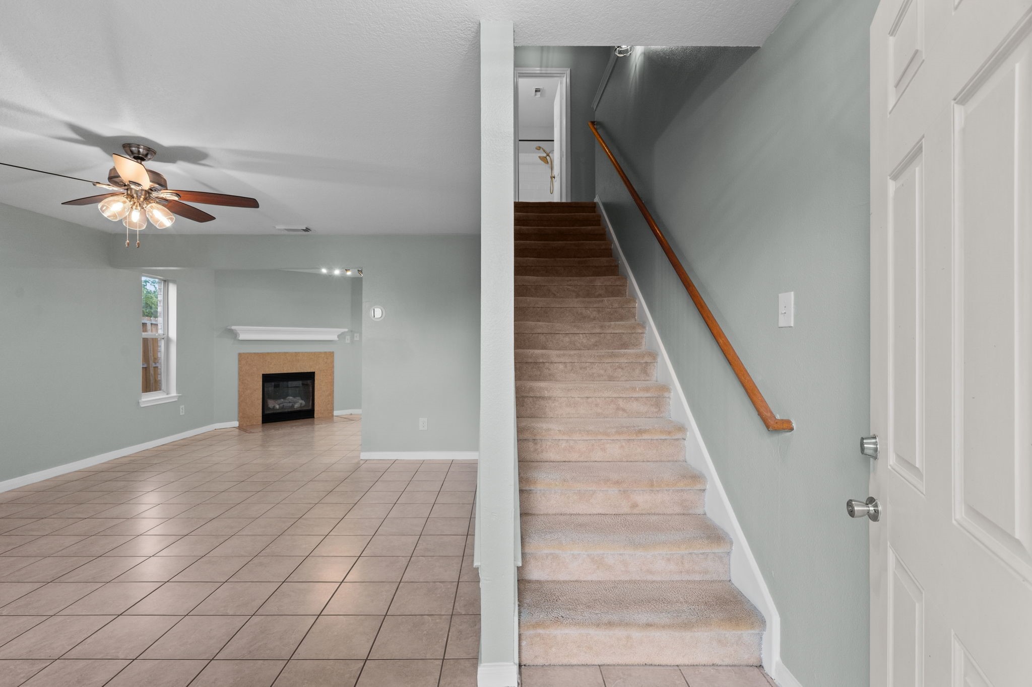 Inviting living space with a cozy fireplace, ceiling fan, and tile flooring, adjacent to a carpeted staircase leading to the upper level.
