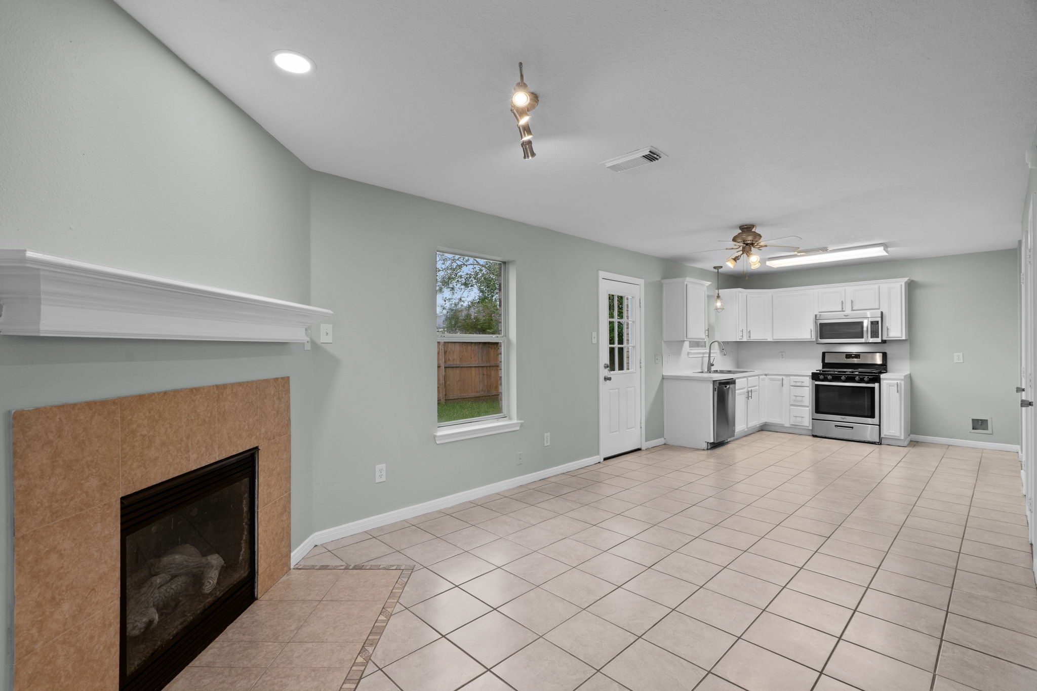 Open-concept living area with a cozy fireplace, light tile flooring, and modern kitchen featuring stainless steel appliances and white cabinetry.