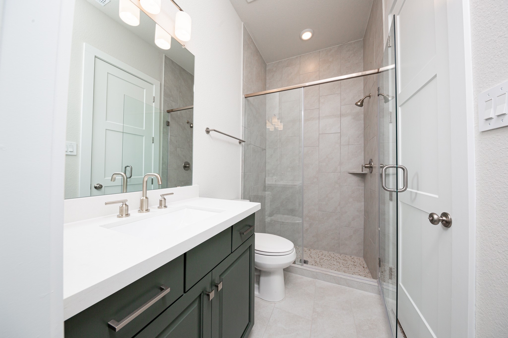 This en-suite bathroom offers stained cabinet topped with quartz countertop and undermount sink, brushed nickel fixtures, and a large walk-in shower with tile surround.