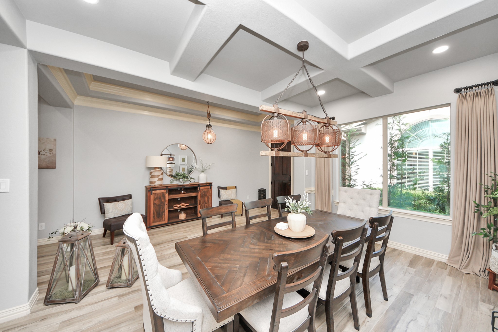 A beautifully designed dining area features a coffered ceiling and elegant light fixtures, with large windows that invite natural light. The space includes a stylish wooden table, comfortable seating, and tasteful decor, creating a warm and inviting atmosphere.
