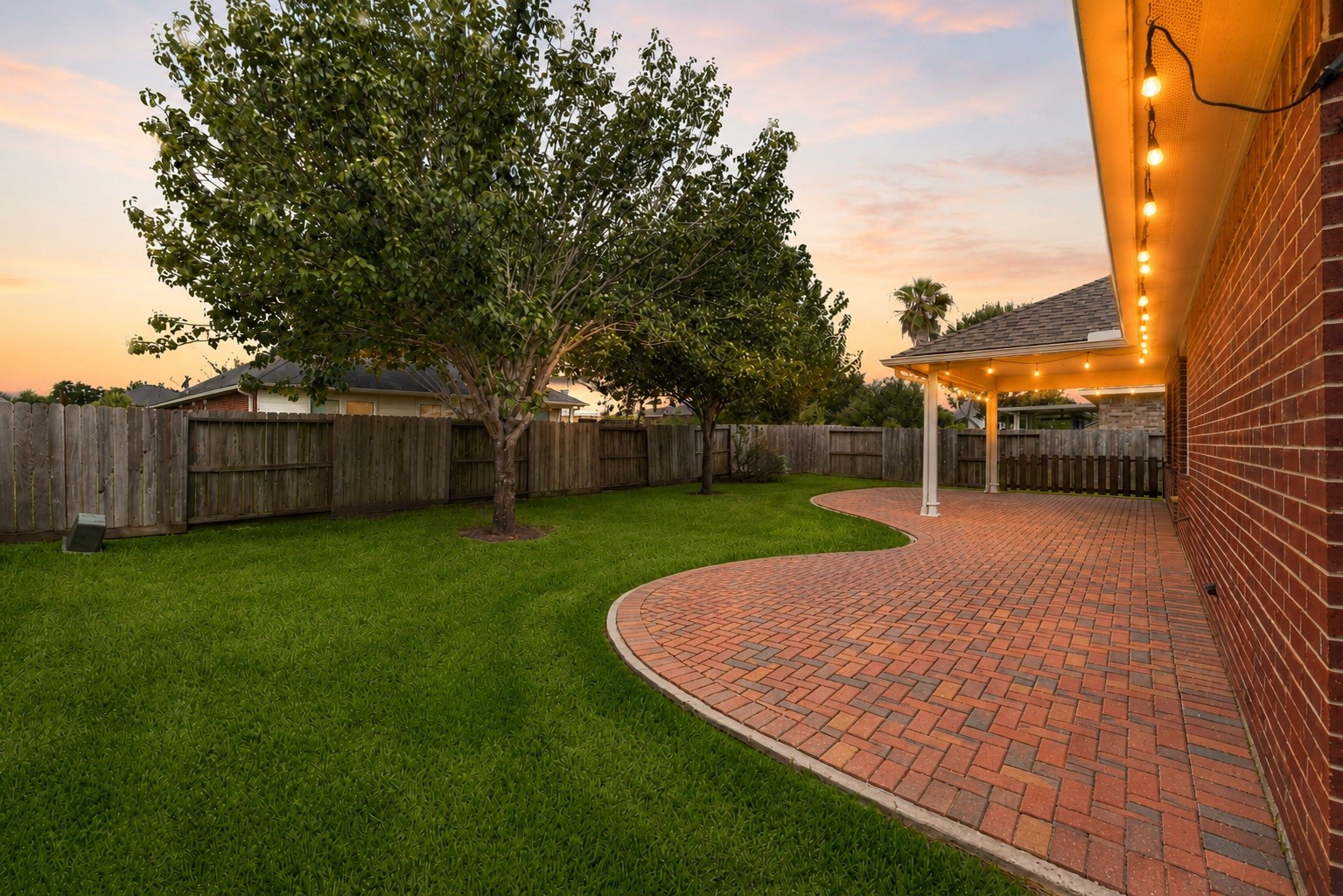 As dusk settles, warm soffit lighting highlights the rear brick elevation and surrounding greenery, lending the backyard a welcoming glow as the day winds down.
