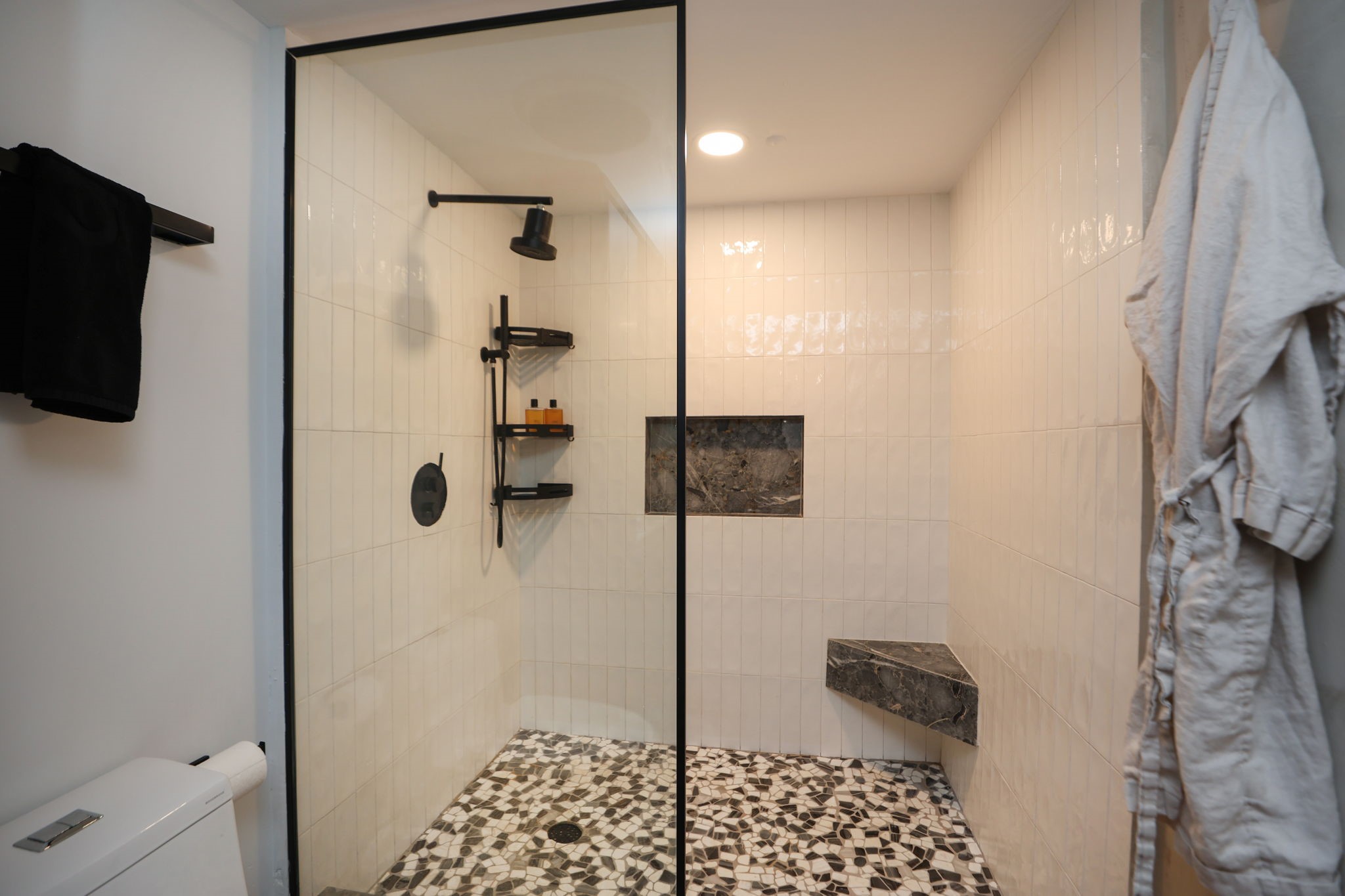 Oversized walk-in shower with a rainfall head, vertical subway tile, pebble-mosaic floor, matte black fixtures, built-in stone bench and a recessed niche for storage.
