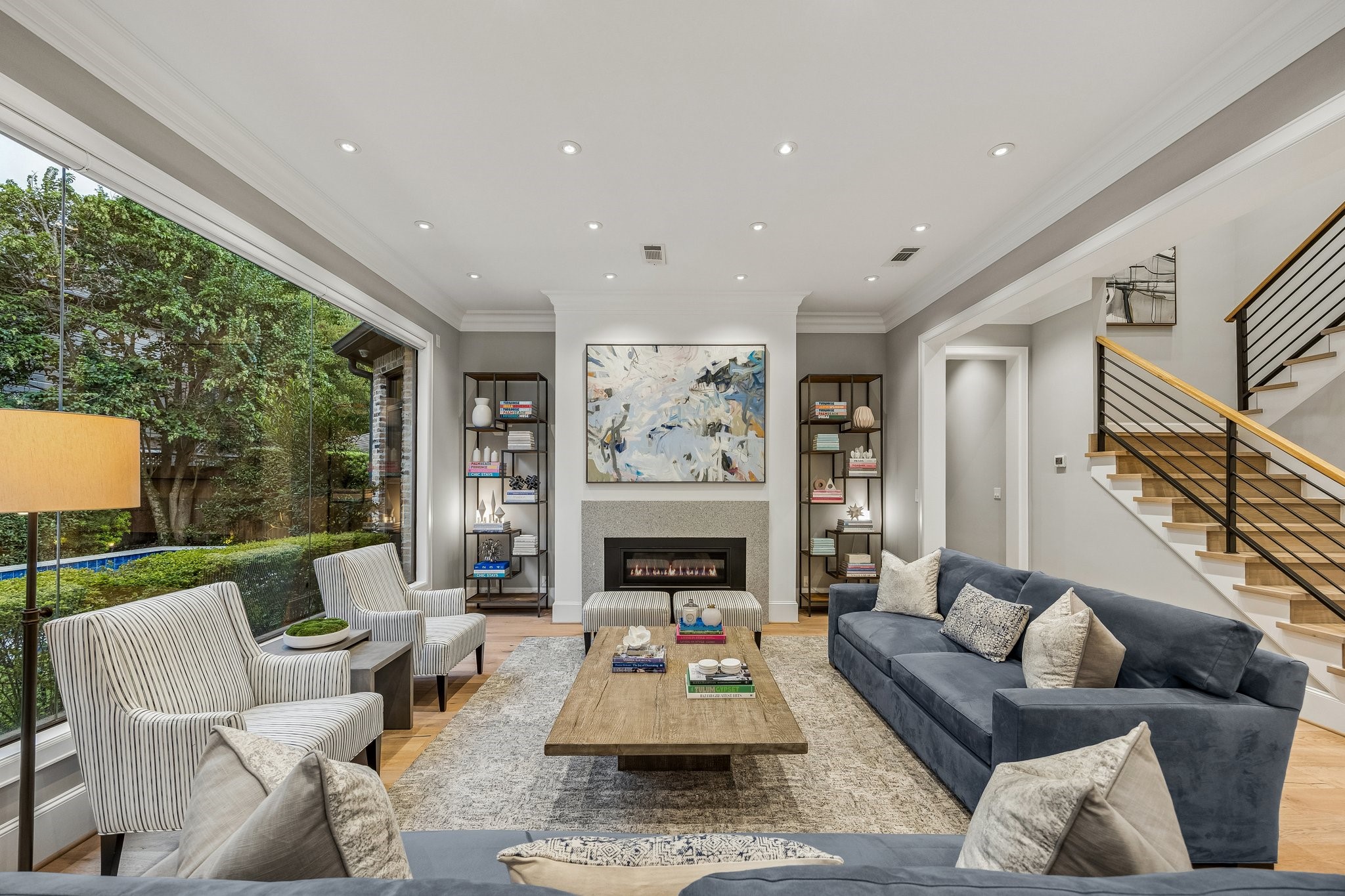 This beautifully appointed living room seamlessly blends contemporary sophistication with timeless architectural elegance. A dramatic floor-to-ceiling picture window serves as the rooms focal point, offering breathtaking views of the lush, meticulously landscaped yard while filling the space with natural light and creating a seamless connection between indoor and outdoor living.
