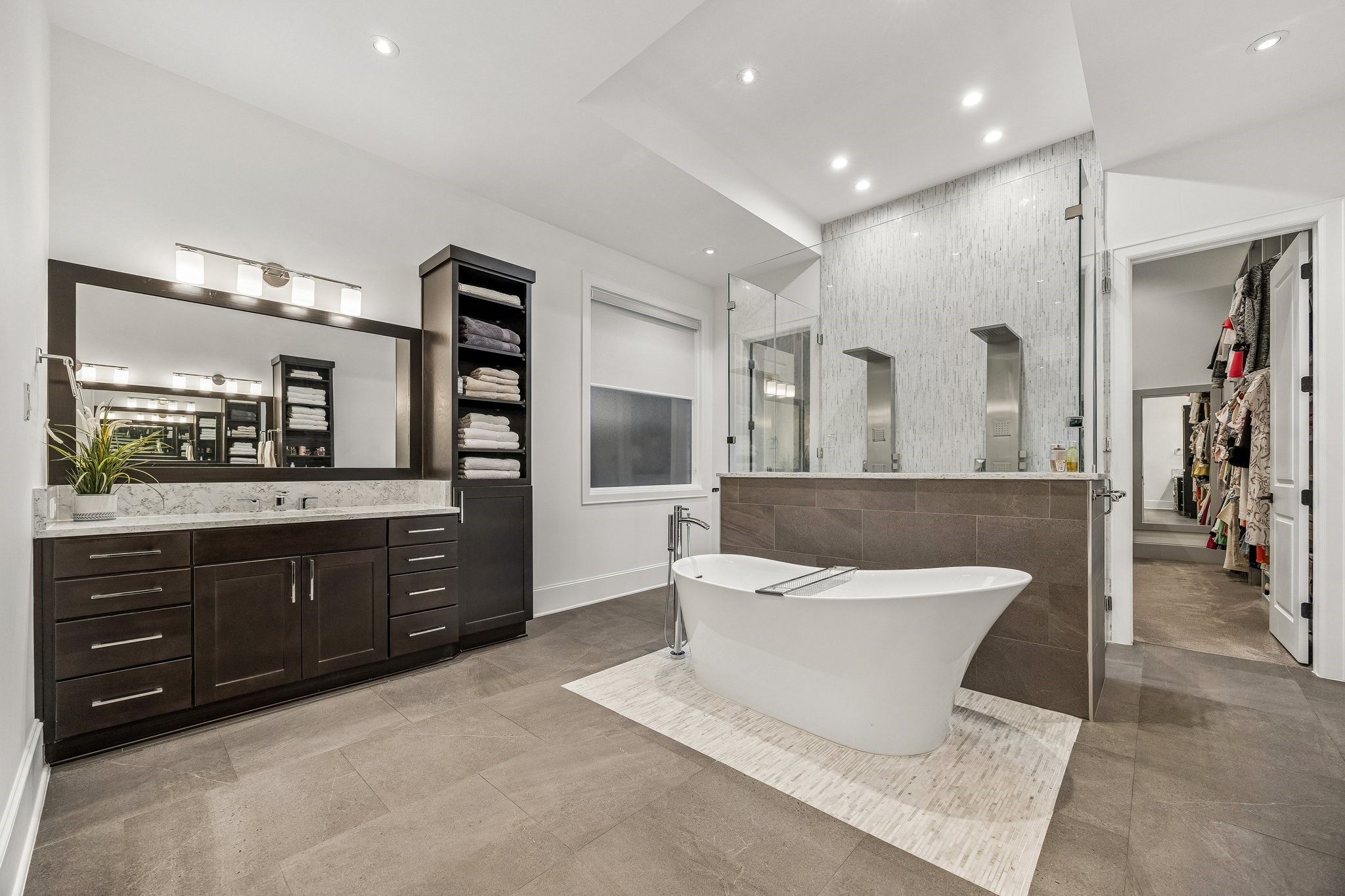 The elegant sculptural soaking tub rests upon a contrasting light-toned mosaic tile platform, beautifully complemented by a sleek, floor-mounted polished chrome tub filler. Beyond the tub, an impressive glass-enclosed walk-in shower features a dramatic floor-to-ceiling mosaic tile accent wall, creating a captivating architectural focal point. Recessed shower niches, contemporary fixtures, and frameless glass panels enhance the clean, modern aesthetic.