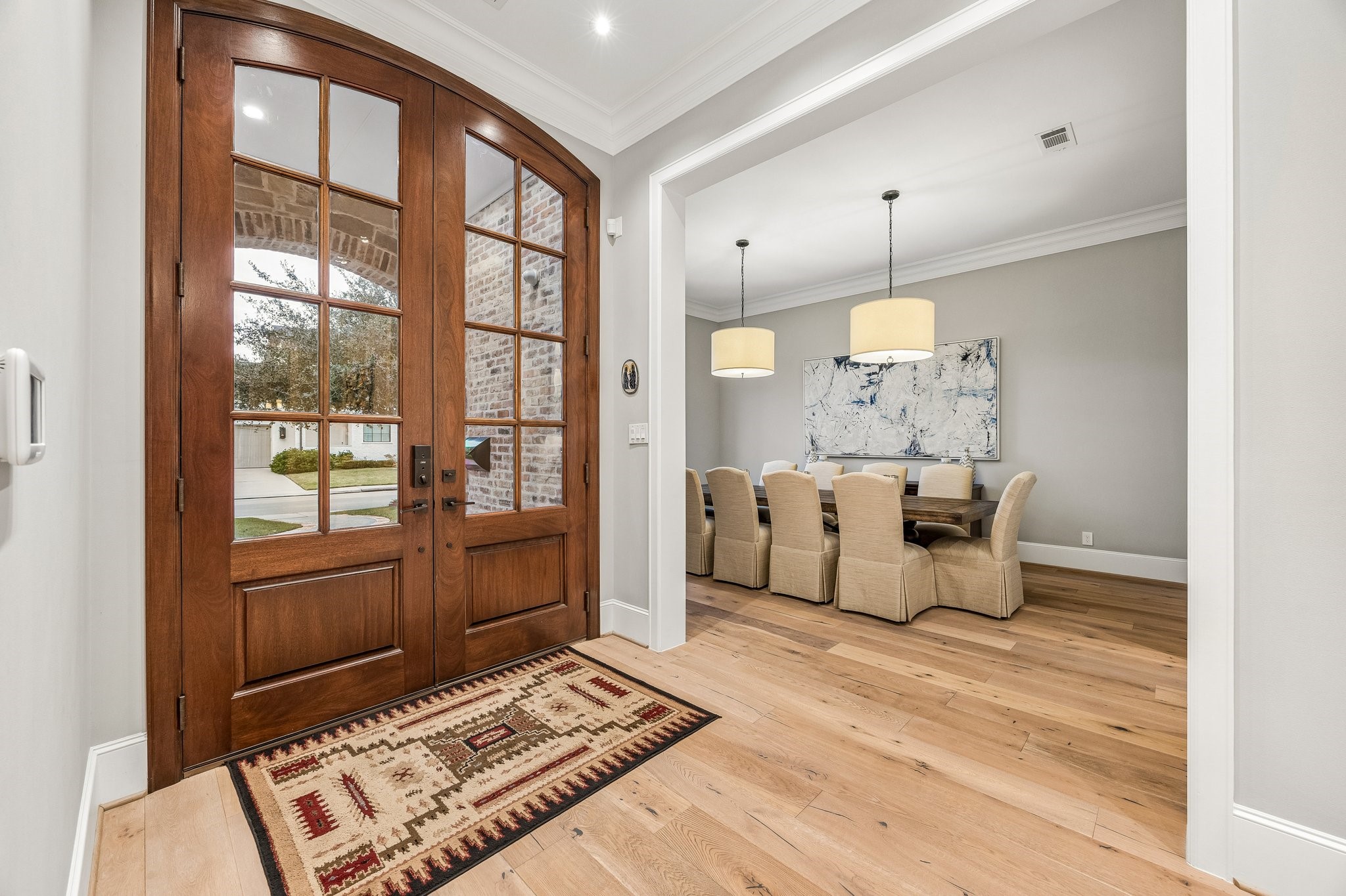 A beautifully appointed entryway welcomes guests with impressive arched, solid wood double doors featuring divided glass panes, creating a warm and inviting first impression. Wide-plank, light-toned hardwood flooring flows seamlessly throughout the foyer and into the elegant formal dining room.