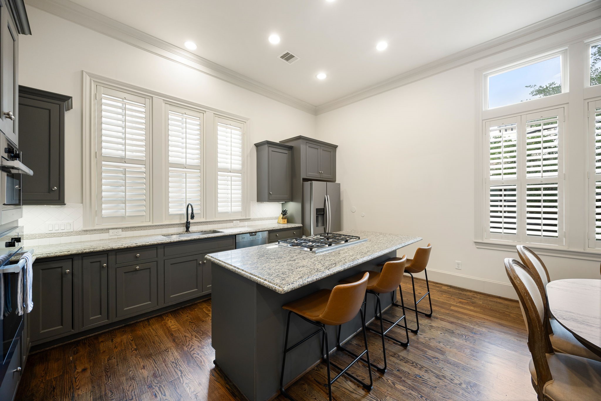This kitchen features elegant custom painted cabinetry, a spacious island with a built-in cooktop, and modern stainless steel appliances. Bright and airy, it has large windows with shutters and warm hardwood floors, creating a welcoming atmosphere.
