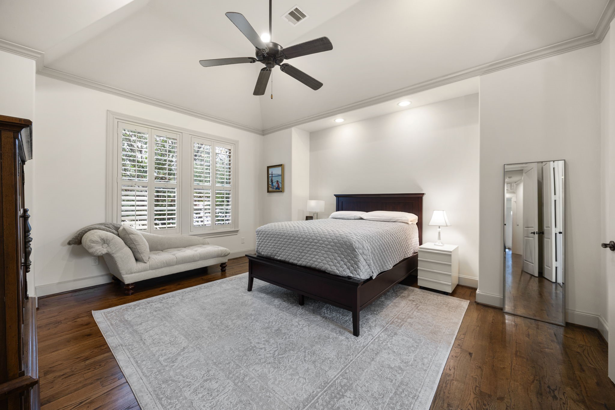 This spacious primary bedroom features elevated finishes such as a vaulted ceiling, crown moulding, hardwood floors, and shuttered windows.  The neutral tones create an elegant place to retreat.