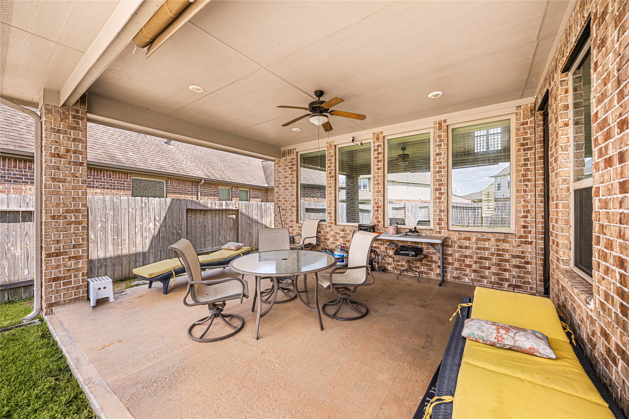 Spacious covered patio with ceiling fan, perfect for outdoor entertaining, featuring comfortable seating and a dining set, surrounded by brick accents and a fenced yard for privacy.