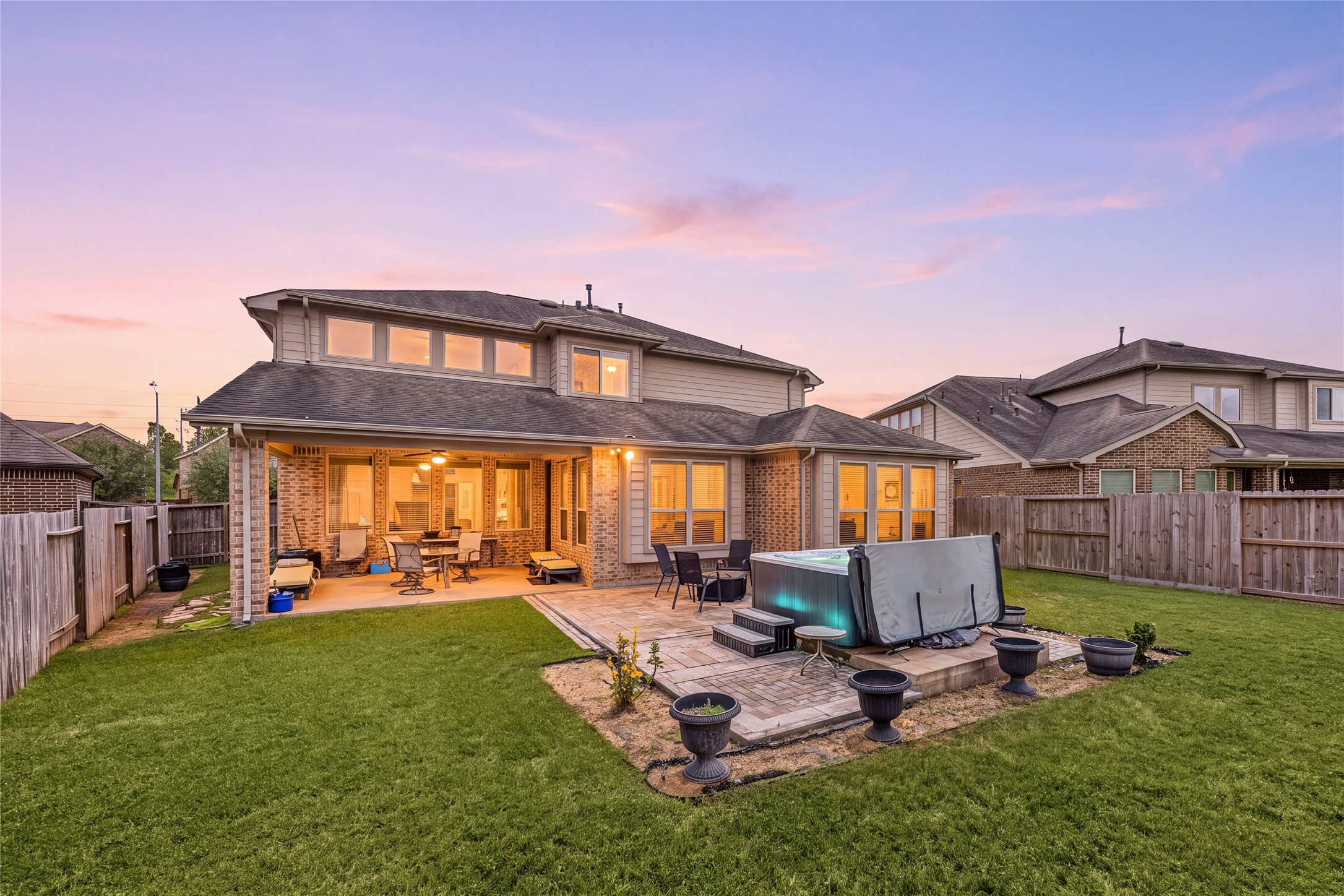 Spacious backyard oasis featuring a large patio with a covered seating area, perfect for outdoor entertaining, and a hot tub for relaxation, all set against a beautiful sunset backdrop.