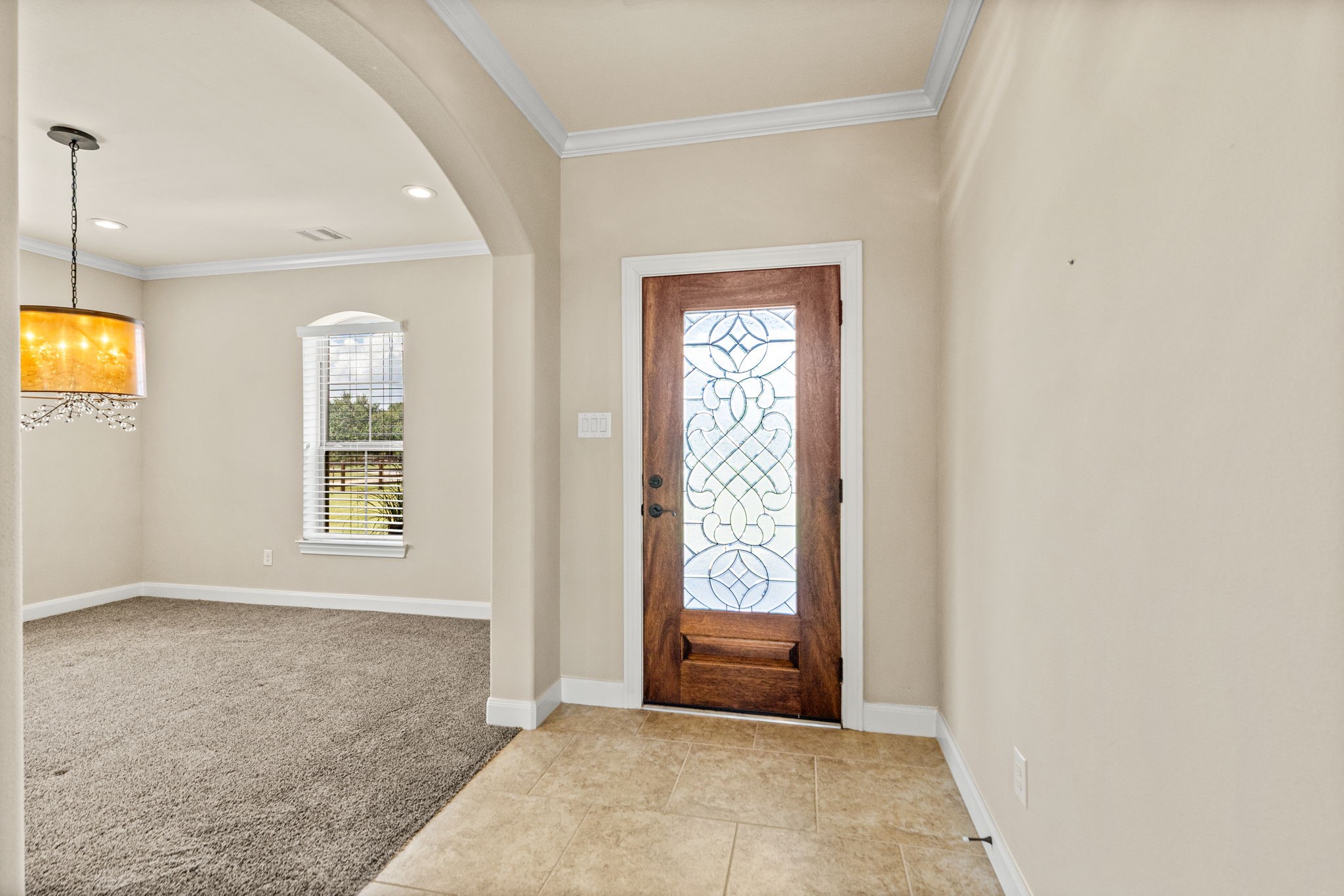 Elegant entryway featuring a beautifully crafted wooden door with intricate glass detailing, leading into a spacious carpeted living area with stylish lighting.