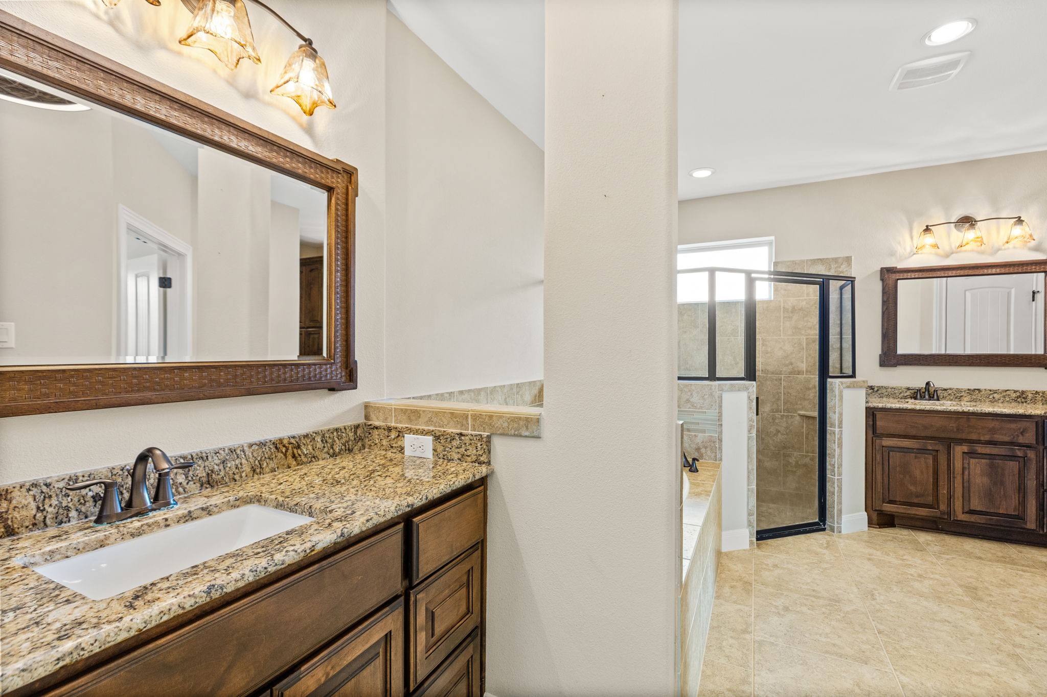Elegant bathroom featuring dual granite vanities, stylish wooden cabinetry, and a spacious walk-in shower with modern tile design.