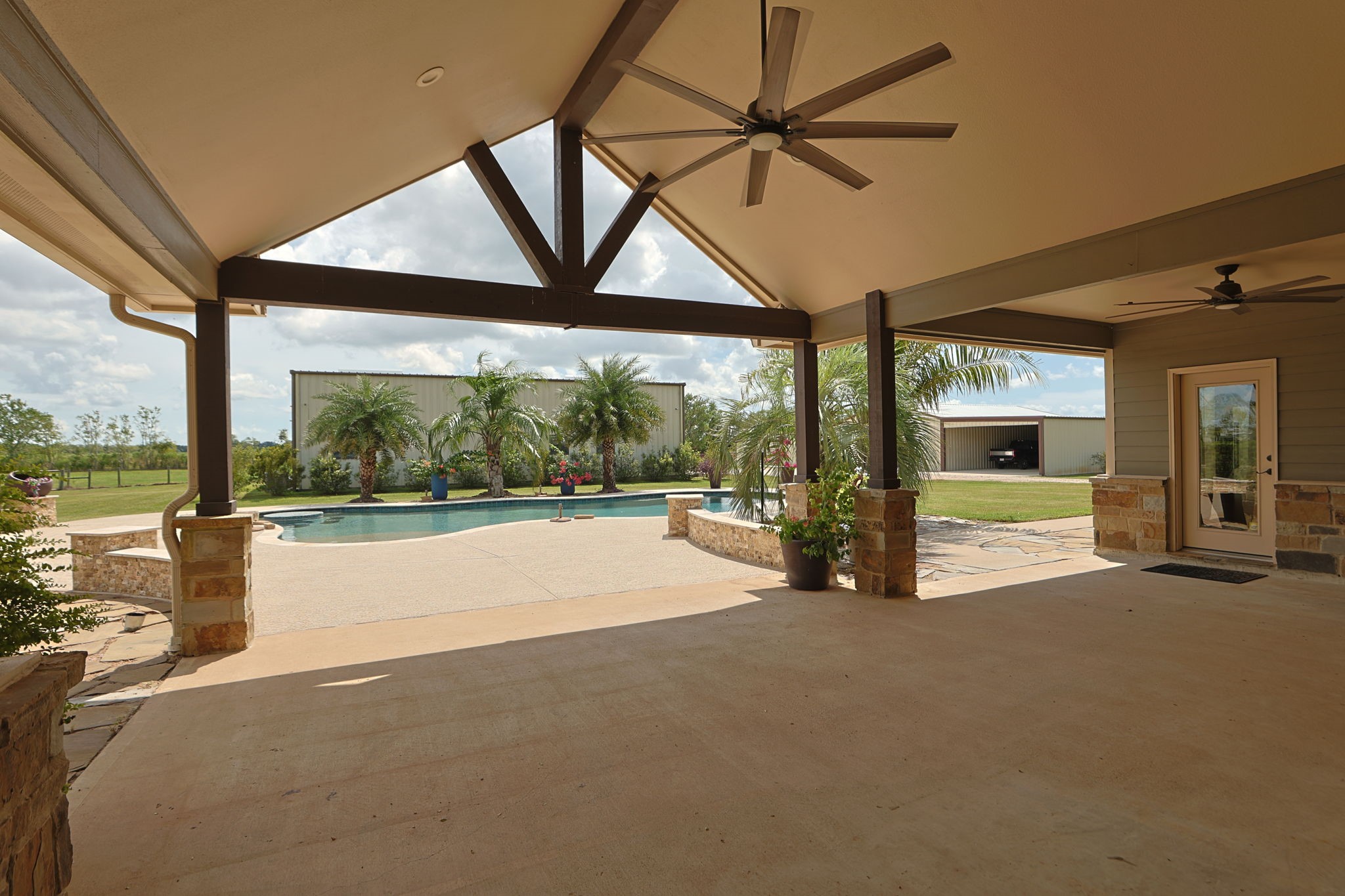 Expansive covered patio with ceiling fans overlooks a sparkling pool surrounded by lush palm trees, creating a perfect outdoor oasis.