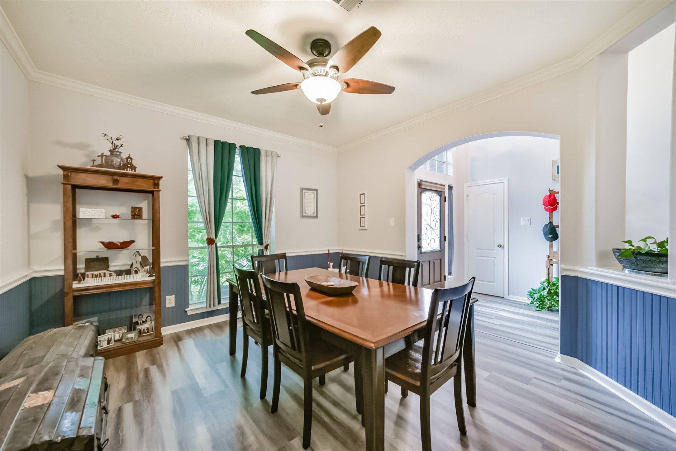 This formal dining room is defined by elegant crown and chair molding, with Beadboard accenting the walls. A ceiling fan with an integrated light fixture hangs overhead, while curtains frame the large window.