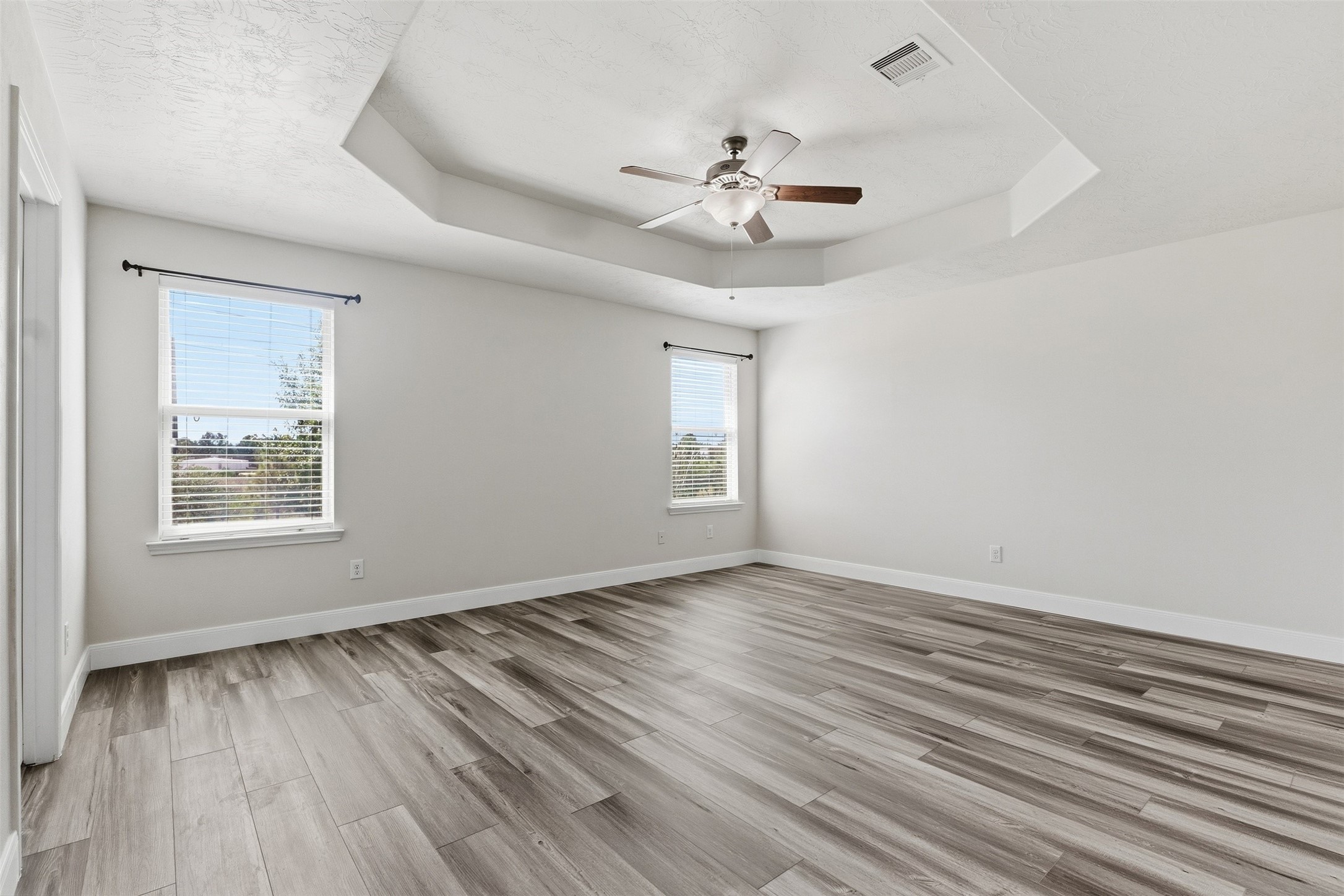 Sizable primary bedroom featuring wood-look flooring, neutral finishes, and a raised tray ceiling with a ceiling fan and light fixture. Multiple windows provide abundant natural light, while the generous room dimensions offer ample space for a variety of bedroom furniture arrangements.