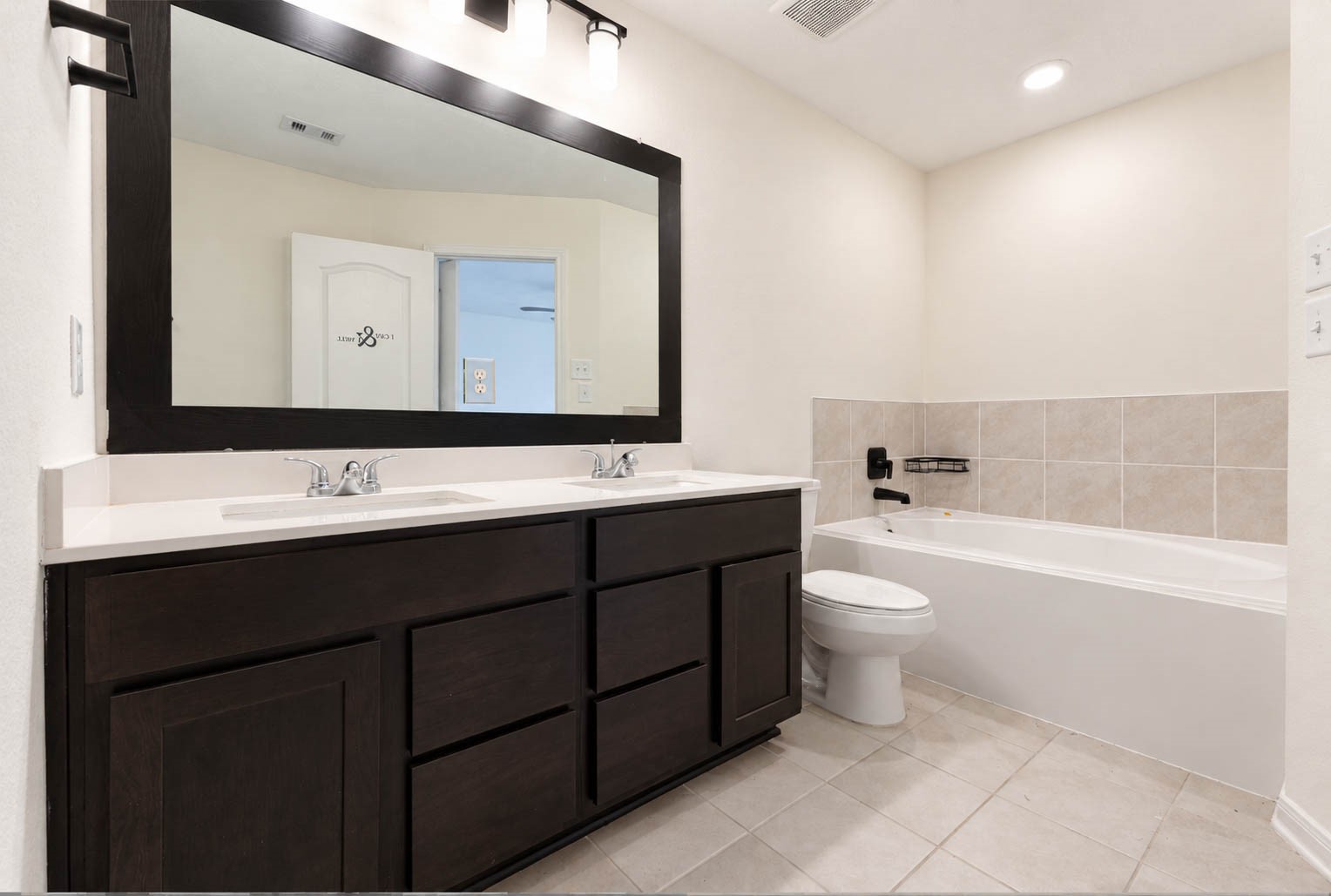 The beautifully appointed primary ensuite bath showcases an expansive double-sink vanity with rich espresso-toned cabinetry, generous countertop space, an oversized framed mirror, & stylish contemporary lighting. A luxurious soaking tub with a coordinating tile surround provides the perfect place to relax & unwind.