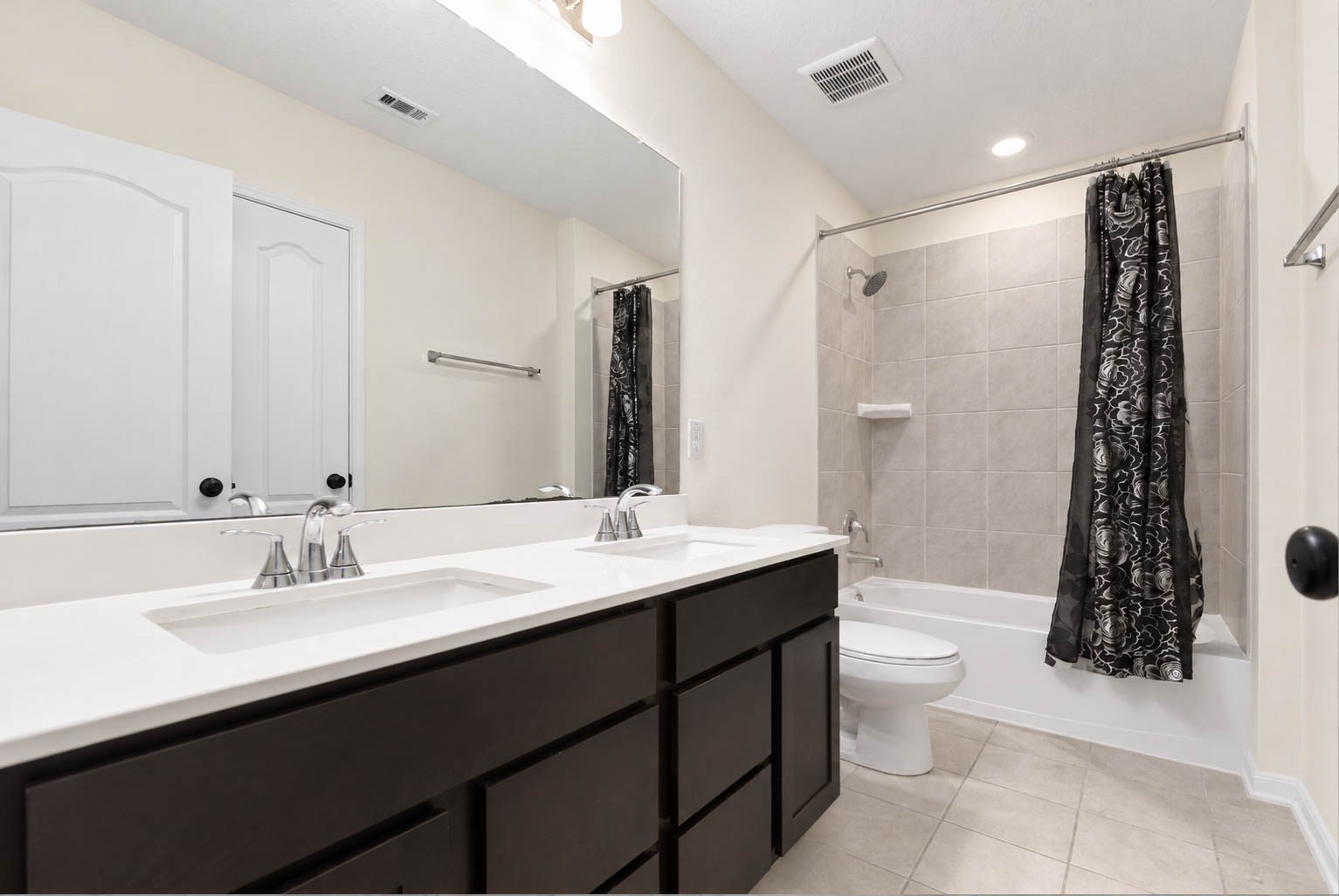 The spacious secondary bathroom features a generous double-sink vanity with crisp white countertops, rich espresso-toned cabinetry, & a a large mirror. Stylish brushed-nickel fixtures, overhead vanity lighting, neutral tile flooring, & a tub/shower combination with coordinating tile surround complete this space.
