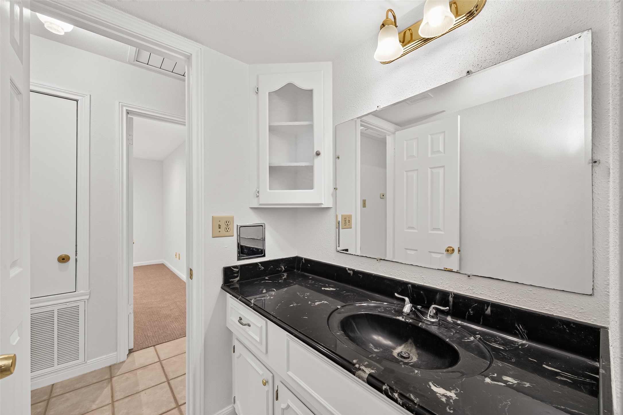 Well-appointed secondary bathroom featuring a striking black marble-look vanity countertop, white cabinetry, a spacious mirror, and decorative lighting. A built-in glass-front cabinet provides additional storage, while neutral tile flooring completes the space.
