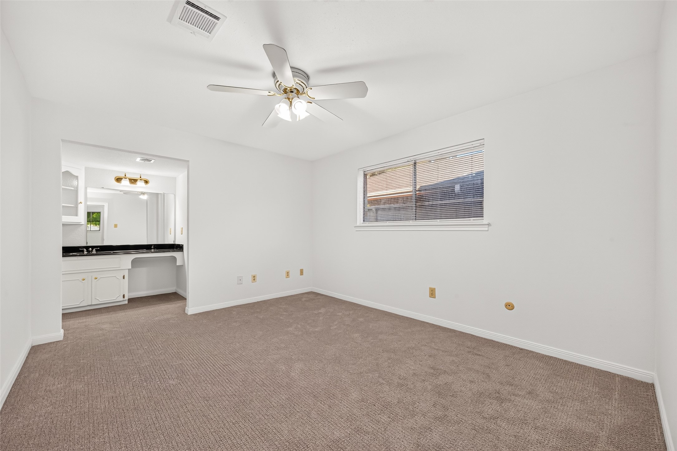 Spacious primary suite featuring fresh neutral paint, plush carpeting, and a ceiling fan. A large window welcomes natural light, creating a bright and relaxing atmosphere. The suite offers convenient access to a private vanity area with generous counter space and built-in cabinetry.