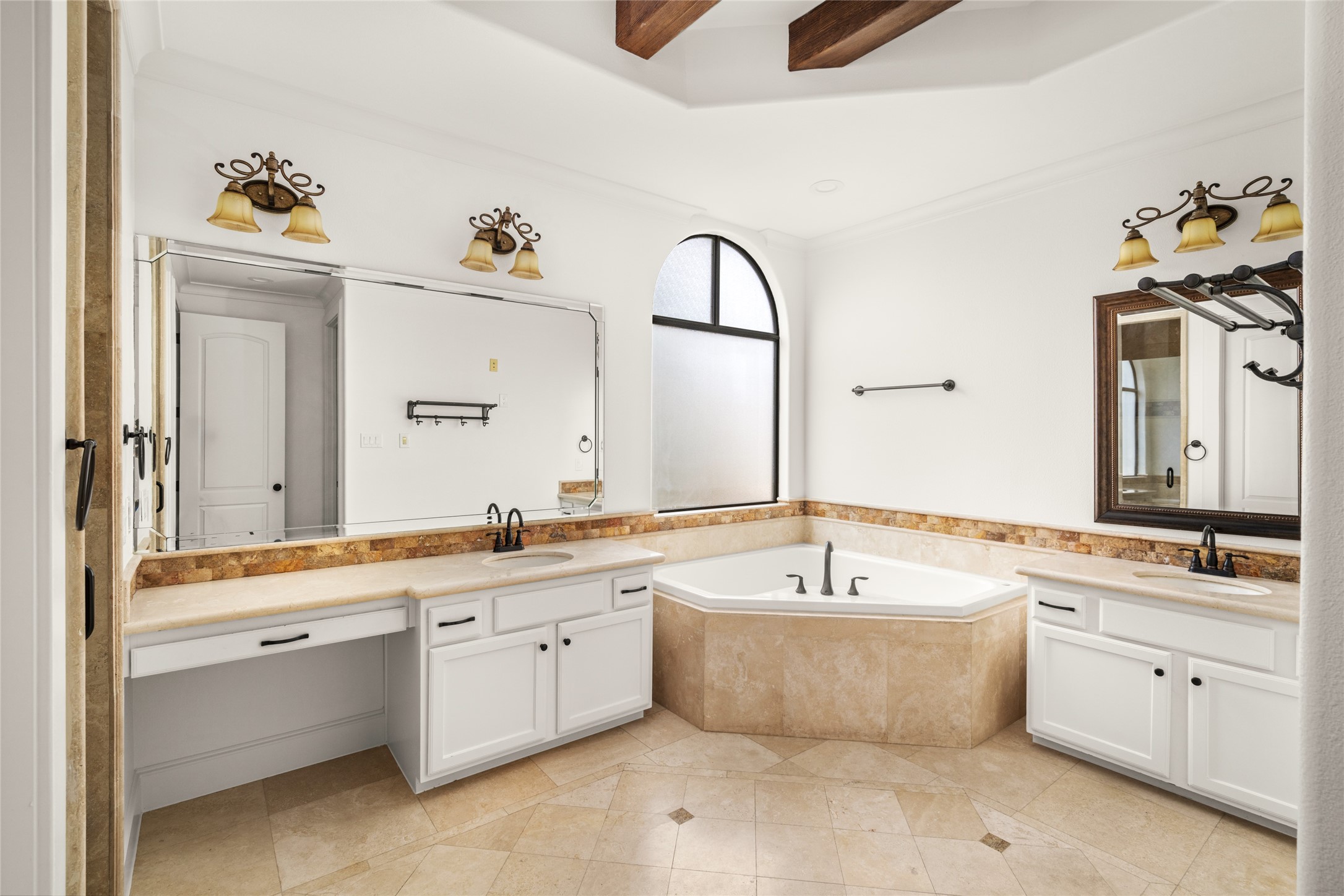 Spacious primary bathroom featuring dual vanities, white cabinetry, a corner soaking tub, and elegant stone tile finishes.