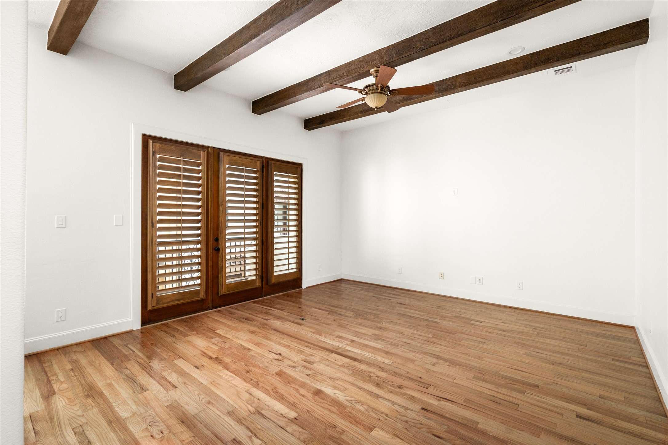 Spacious second-floor living room featuring refinished oak hardwood floors, exposed wood ceiling beams, a ceiling fan, and French doors with plantation shutters.