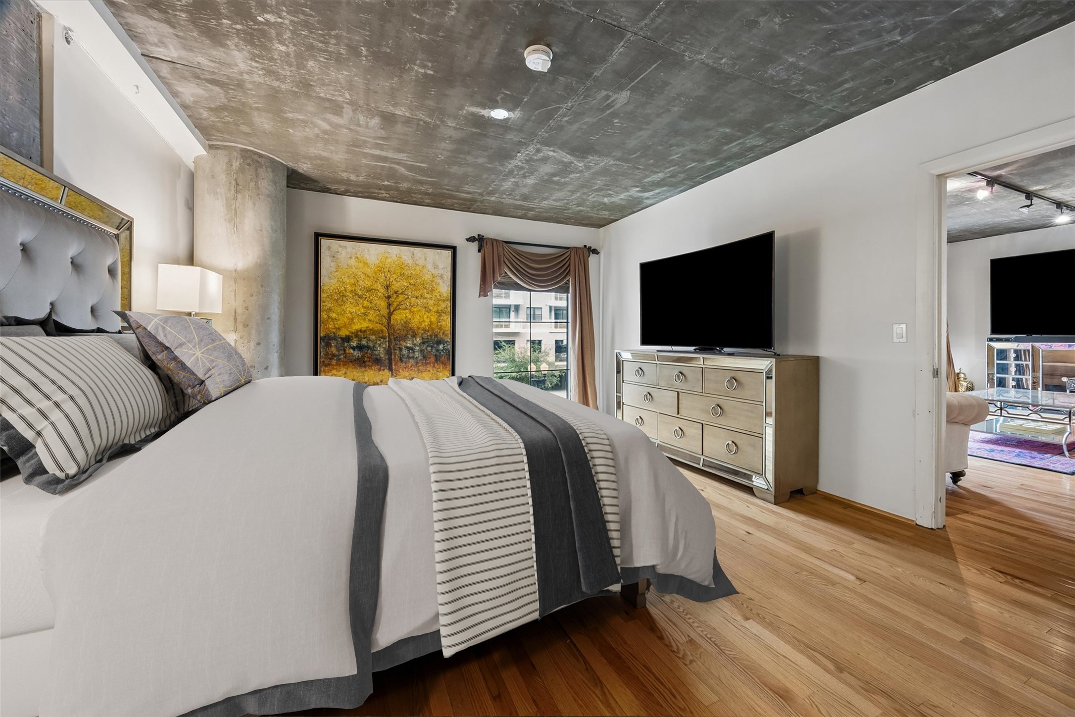 Another perspective of the primary bedroom showcases its generous proportions, hardwood flooring, and distinctive exposed concrete ceiling. The layout comfortably accommodates a large bed and additional furnishings while maintaining an open, inviting feel.