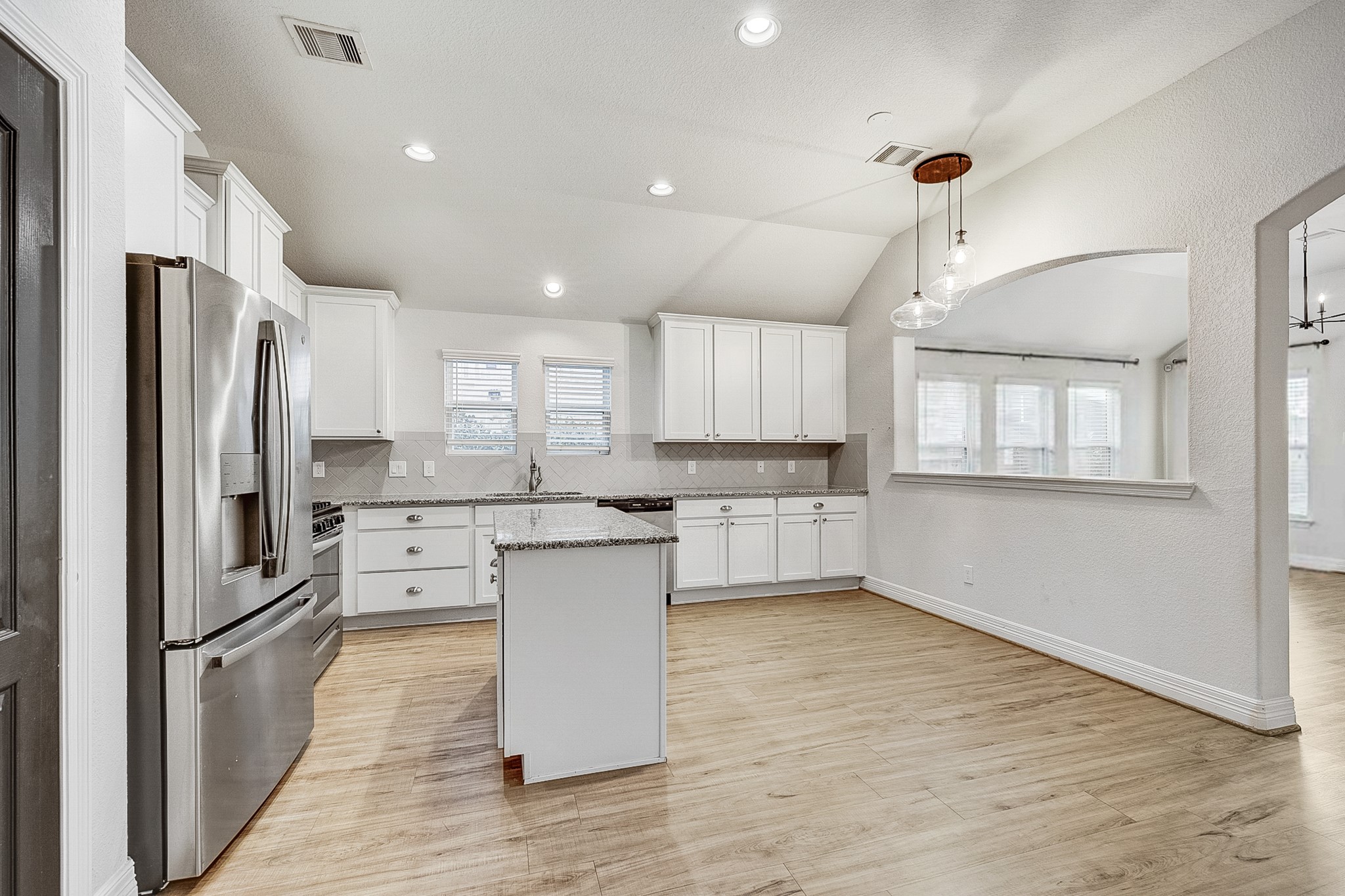 Spacious kitchen featuring modern stainless steel appliances, elegant white cabinetry, and a central island, all set on warm wood flooring and illuminated by stylish pendant lighting.