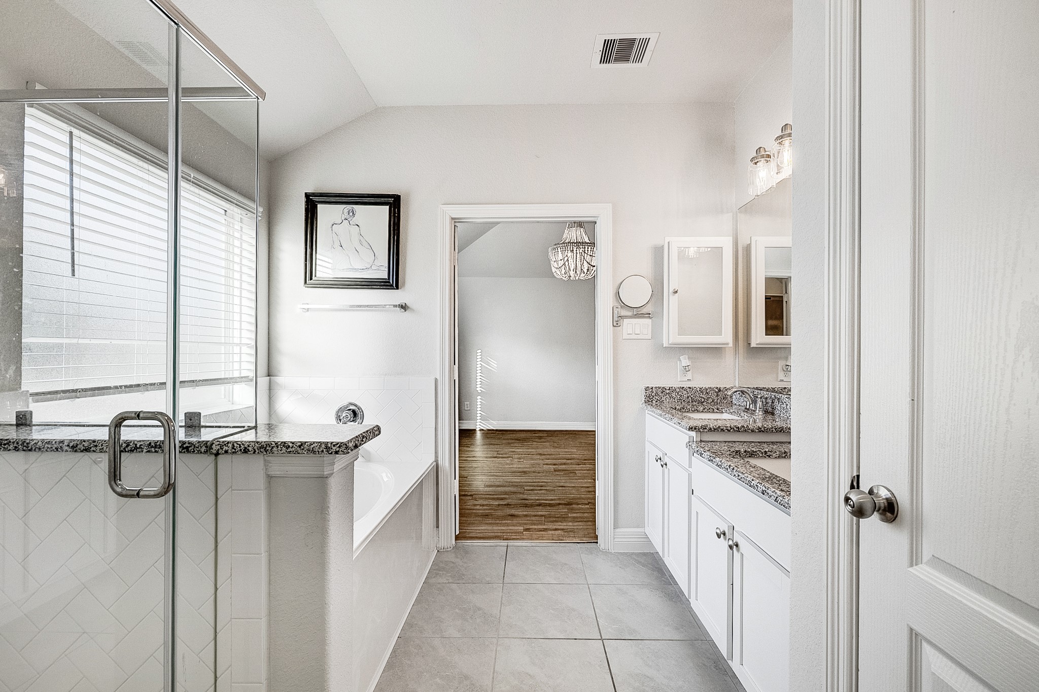 This elegant bathroom features a spacious glass-enclosed shower, a luxurious soaking tub, and dual vanities with granite countertops, leading to a bright room with hardwood flooring.