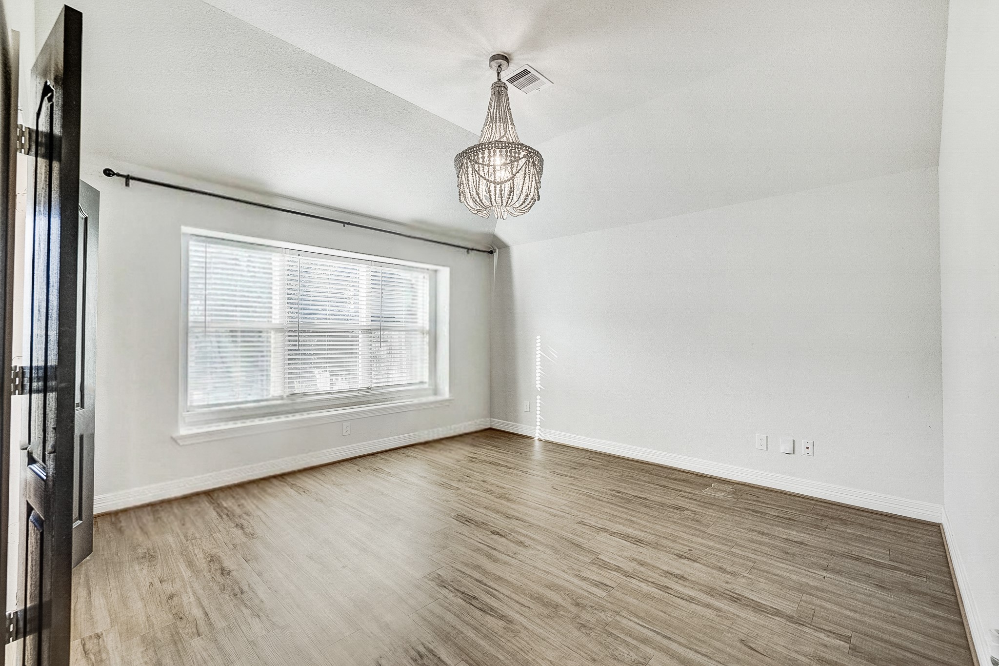 Bright and spacious room featuring elegant wood flooring, a large window with abundant natural light, and a stylish chandelier accentuating the high ceiling.