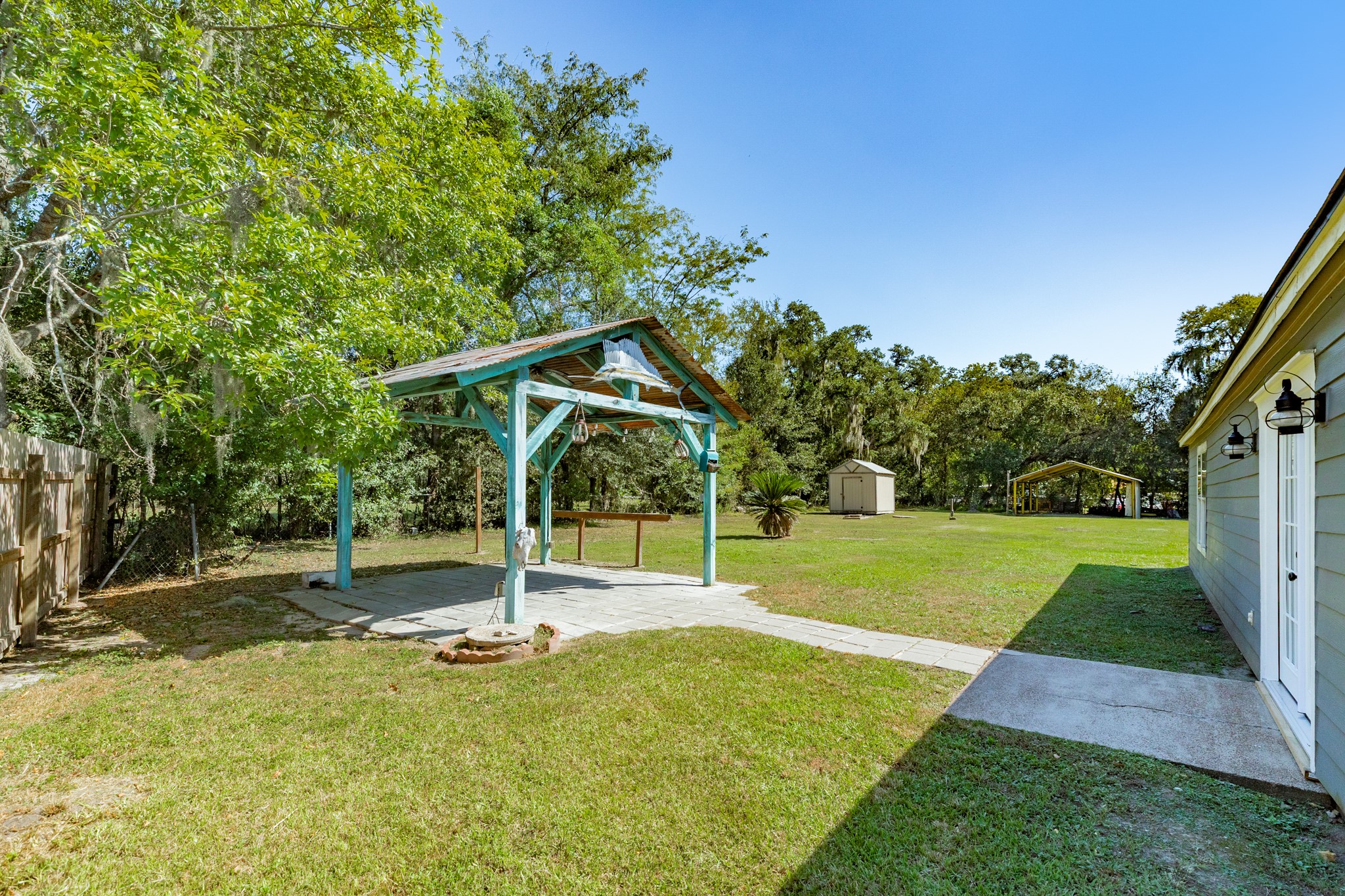 Spacious backyard offers a covered pavilion connected by a paved walkway, surrounded by mature shade trees and open lawn. Additional outdoor structures provide convenient options for storage and covered use.