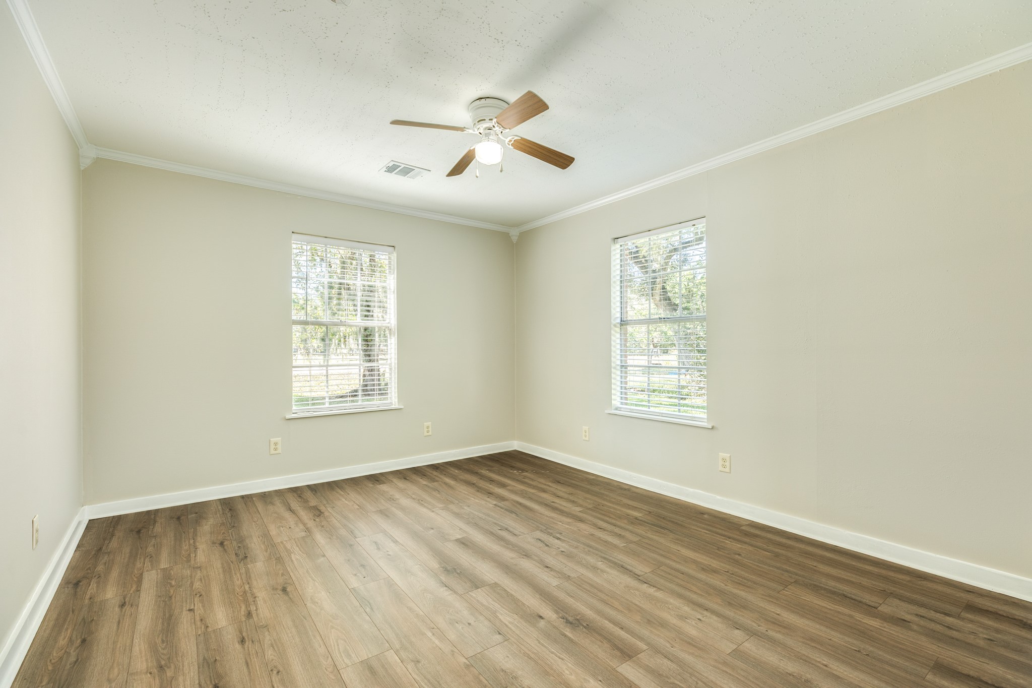 Bright, versatile bedroom showcases attractive wood-look flooring, neutral walls, white trim, and a ceiling fan. Two windows provide natural light and views of the outdoors.