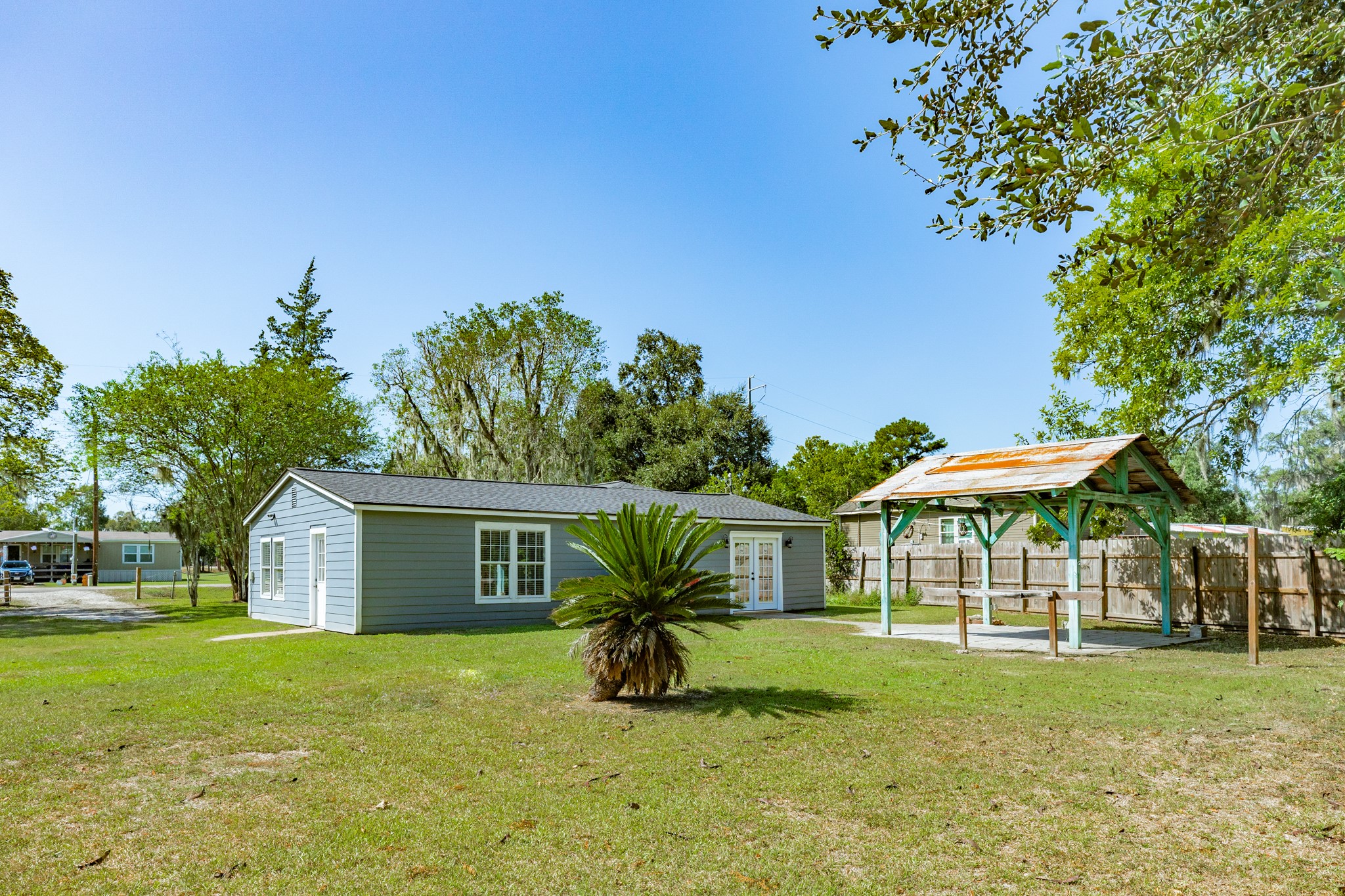 Beautifully open yard features mature landscaping, a decorative palm, and a freestanding covered pavilion with a paved surface. A wonderful setting for outdoor gatherings and enjoying the surrounding greenery.