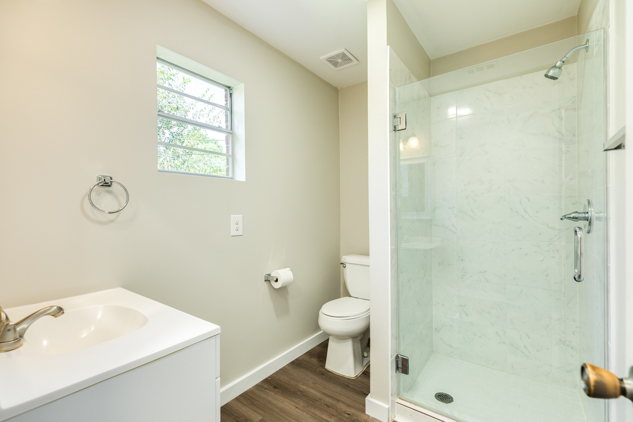 Fresh, neutral bathroom features a glass-enclosed walk-in shower with marble-look wall tile, a white vanity, and wood-look flooring. A window provides natural light and ventilation.