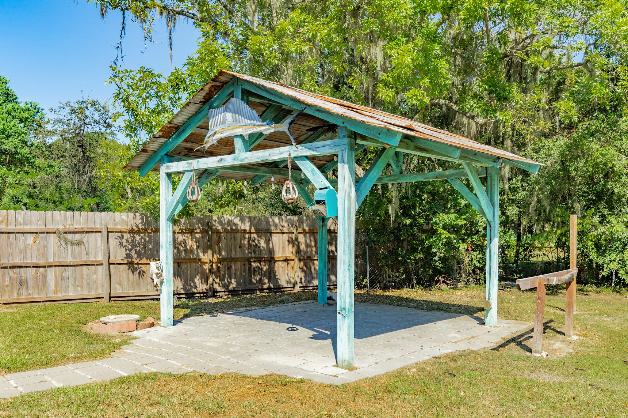 Rustic open-air pavilion features turquoise-painted wooden beams, a metal roof, hanging light fixtures, and a paved foundation. A distinctive outdoor space with room for seating and entertaining.