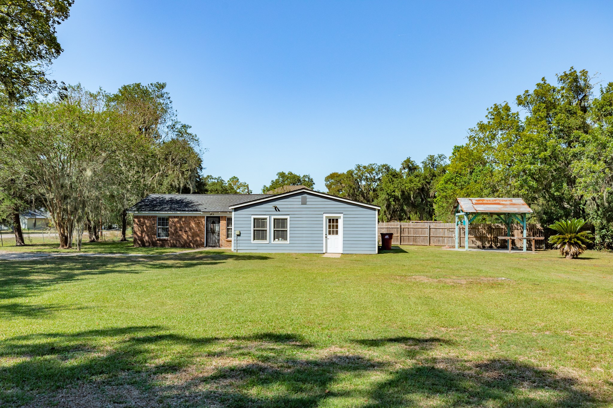 Wide-angle view highlights the home's generous lawn space, mature trees, and open surroundings. A covered outdoor pavilion adds versatility for relaxing or entertaining.