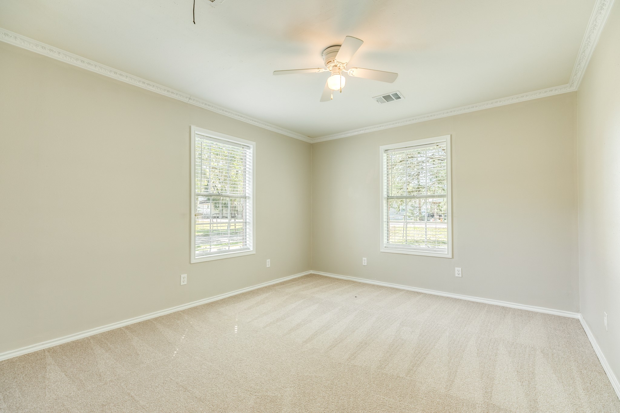 Comfortable bedroom features plush neutral carpeting, decorative crown molding, and a ceiling fan with lighting. Two windows welcome natural light and offer pleasant views of the surrounding greenery.