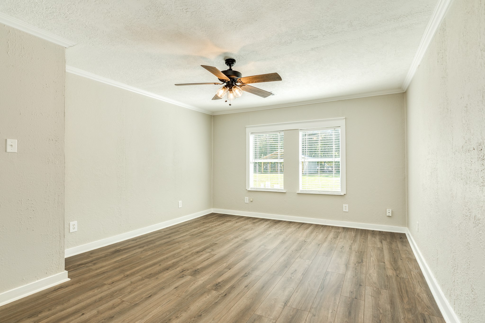 Generously sized bedroom features wood-look flooring, neutral walls, crown molding, and a ceiling fan with lighting. Two windows welcome natural light and offer views of the surrounding greenery.