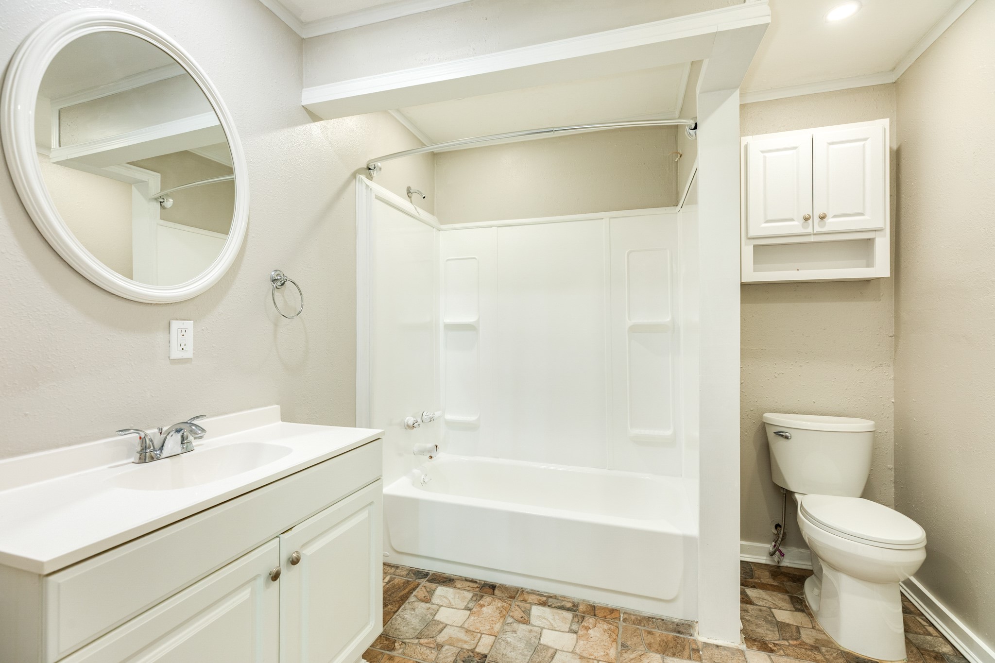 Full bathroom features a white vanity, oval mirror, tub and shower combination with built-in shelving, and additional wall-mounted cabinetry. Decorative stone-look flooring adds warmth and character.