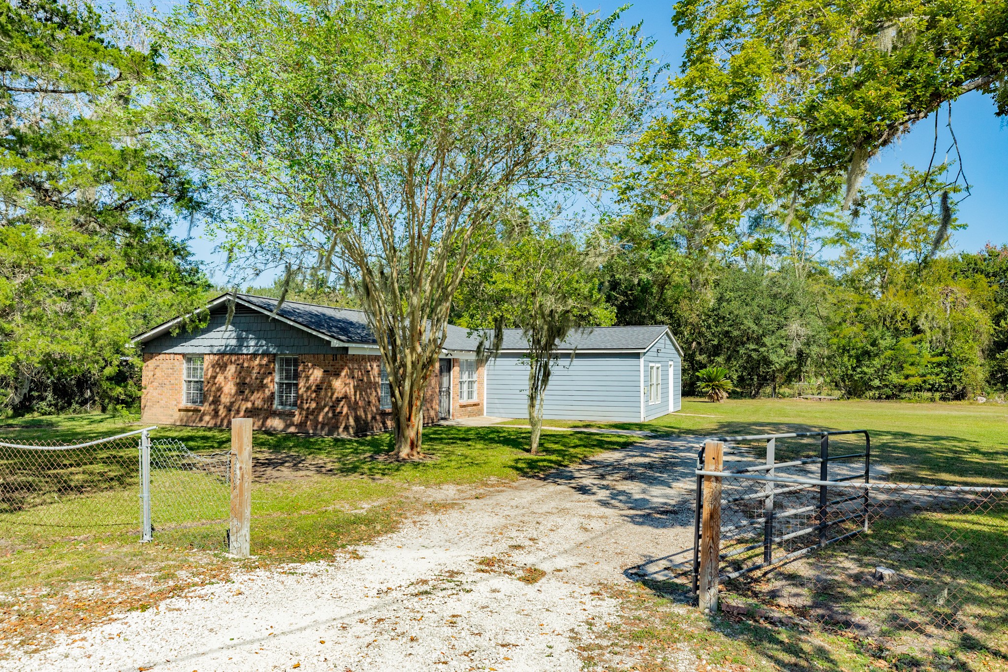 Welcoming single-story home nestled among mature shade trees, featuring a combination of brick and blue-gray siding. A gated entrance and gravel driveway lead to the home, surrounded by expansive green lawn space.