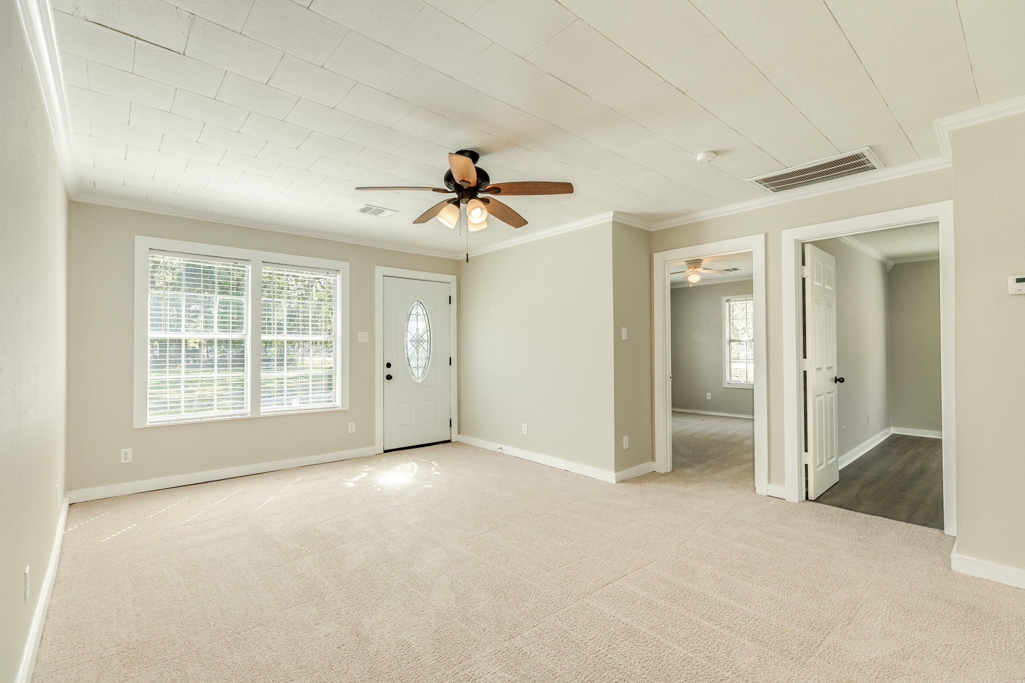 Bright and inviting living room features neutral carpeting, soft wall colors, decorative ceiling panels, and a ceiling fan with lighting. Large windows bring in natural light, while the decorative glass entry door adds character.