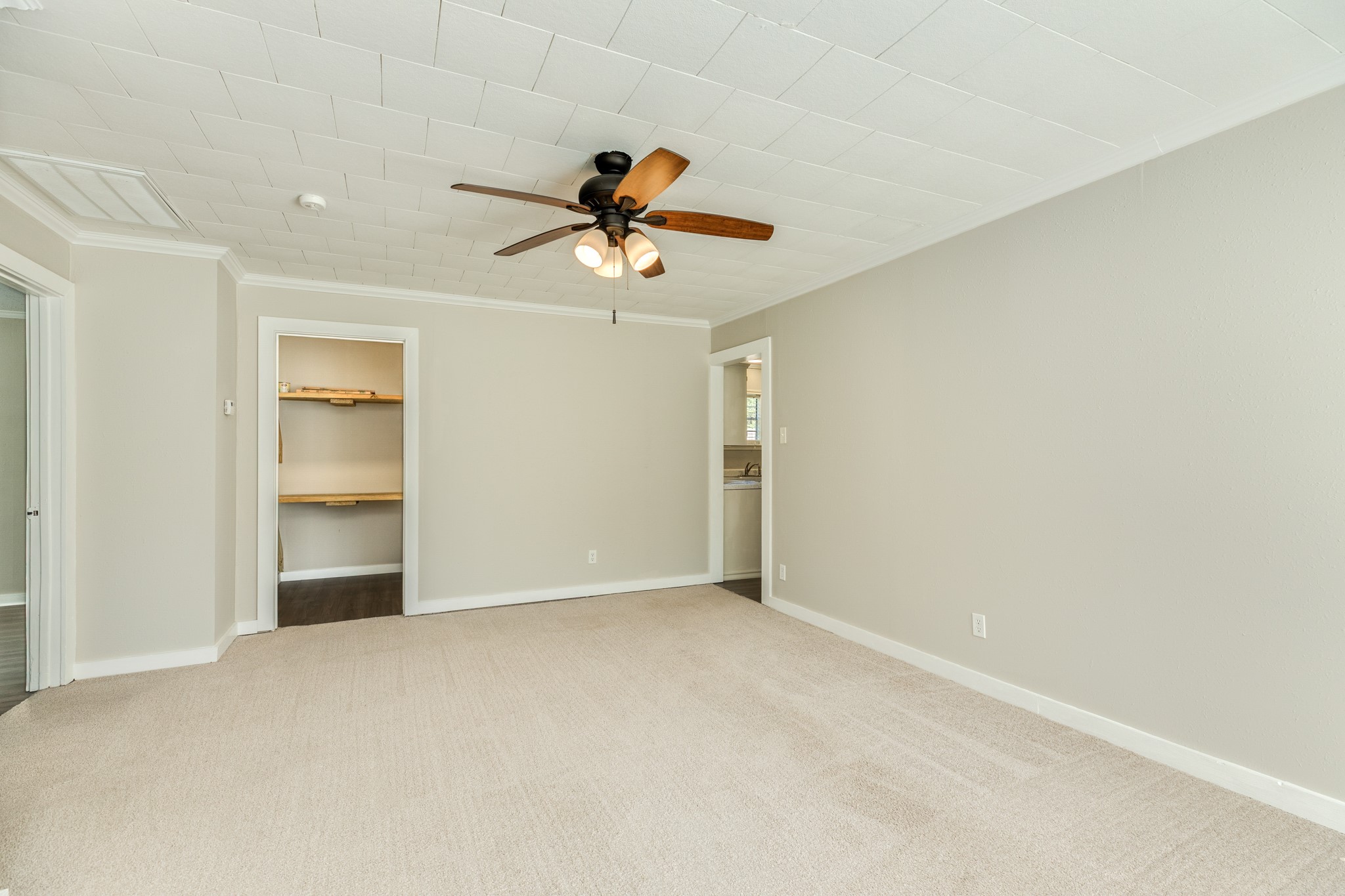 Generously sized living area offers comfortable carpeting, crown molding, and a ceiling fan with warm wood-tone blades. An open doorway provides access to the kitchen, while a recessed shelving area offers convenient storage.