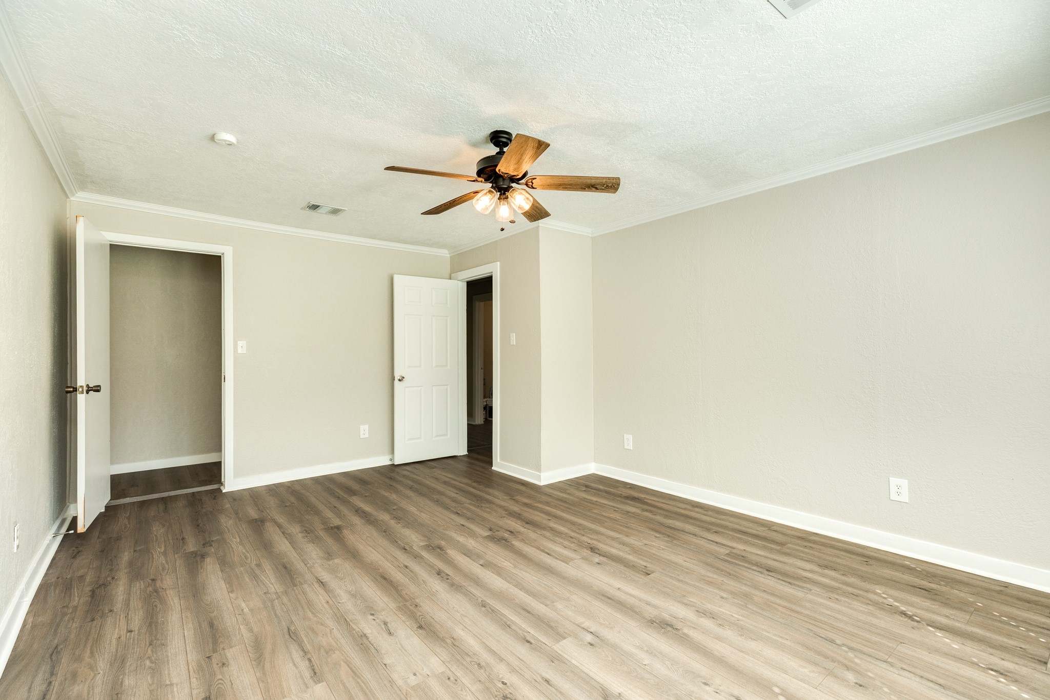 Another view highlights the bedroom's spacious layout, wood-look flooring, and neutral finishes. A ceiling fan with lighting and convenient access to the closet and adjoining hallway complete the space.