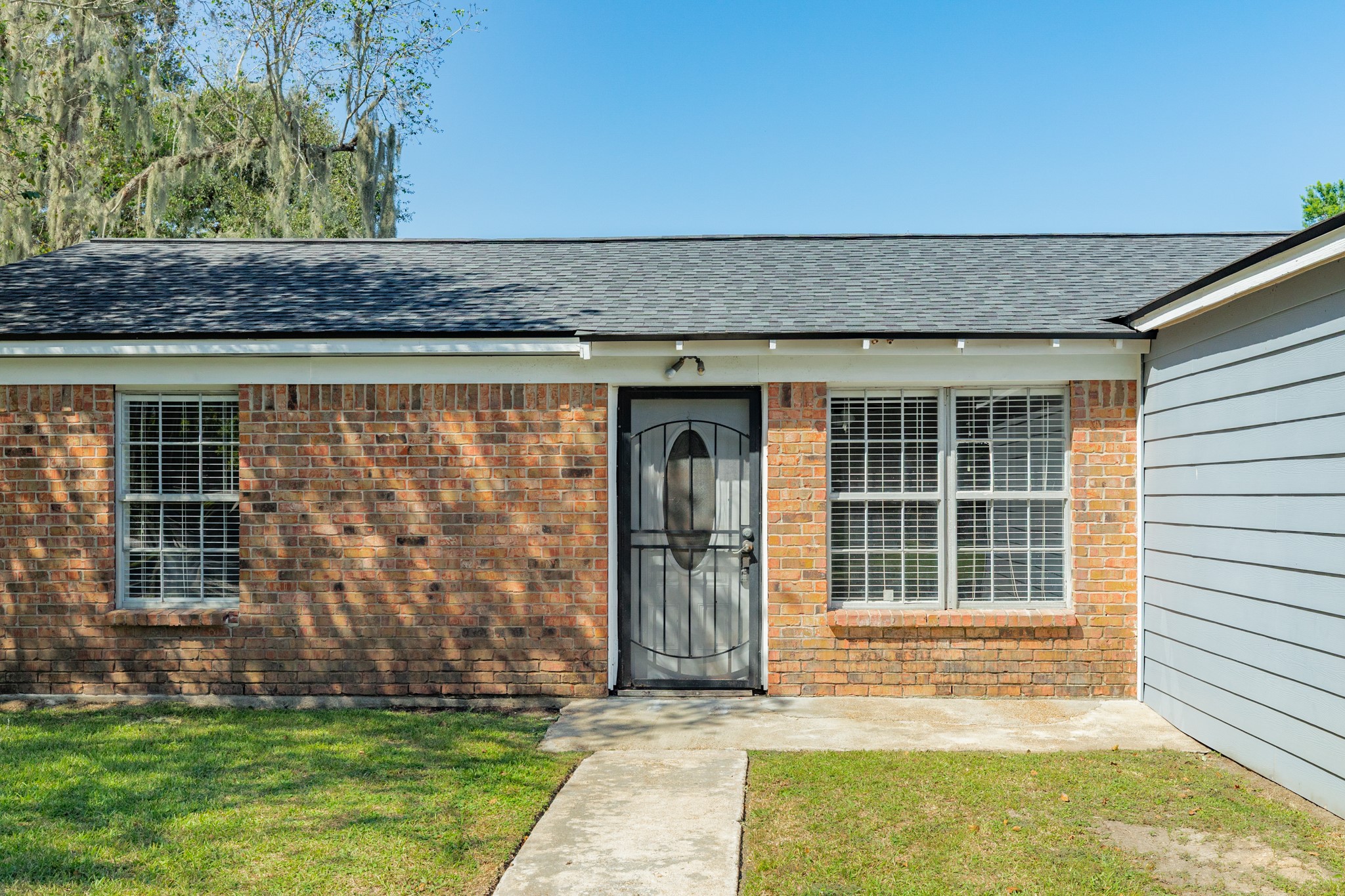 Charming brick faade features a decorative security door, white-trimmed windows, and a concrete walkway. Natural brick tones and mature landscaping add character to the home's entrance.