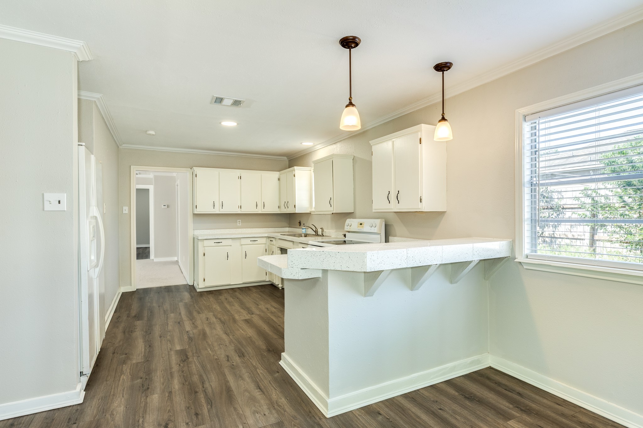 Bright kitchen features white cabinetry, speckled countertops, and a spacious breakfast bar accented by pendant lighting. Wood-look flooring and a large window complement the inviting, functional layout.